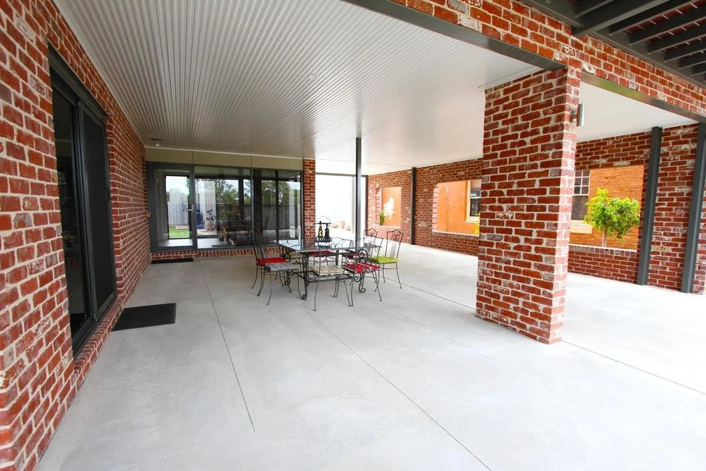 Empty covered outdoor patio with brick columns, concrete floor, metal dining table with chairs, and potted trees in the background.