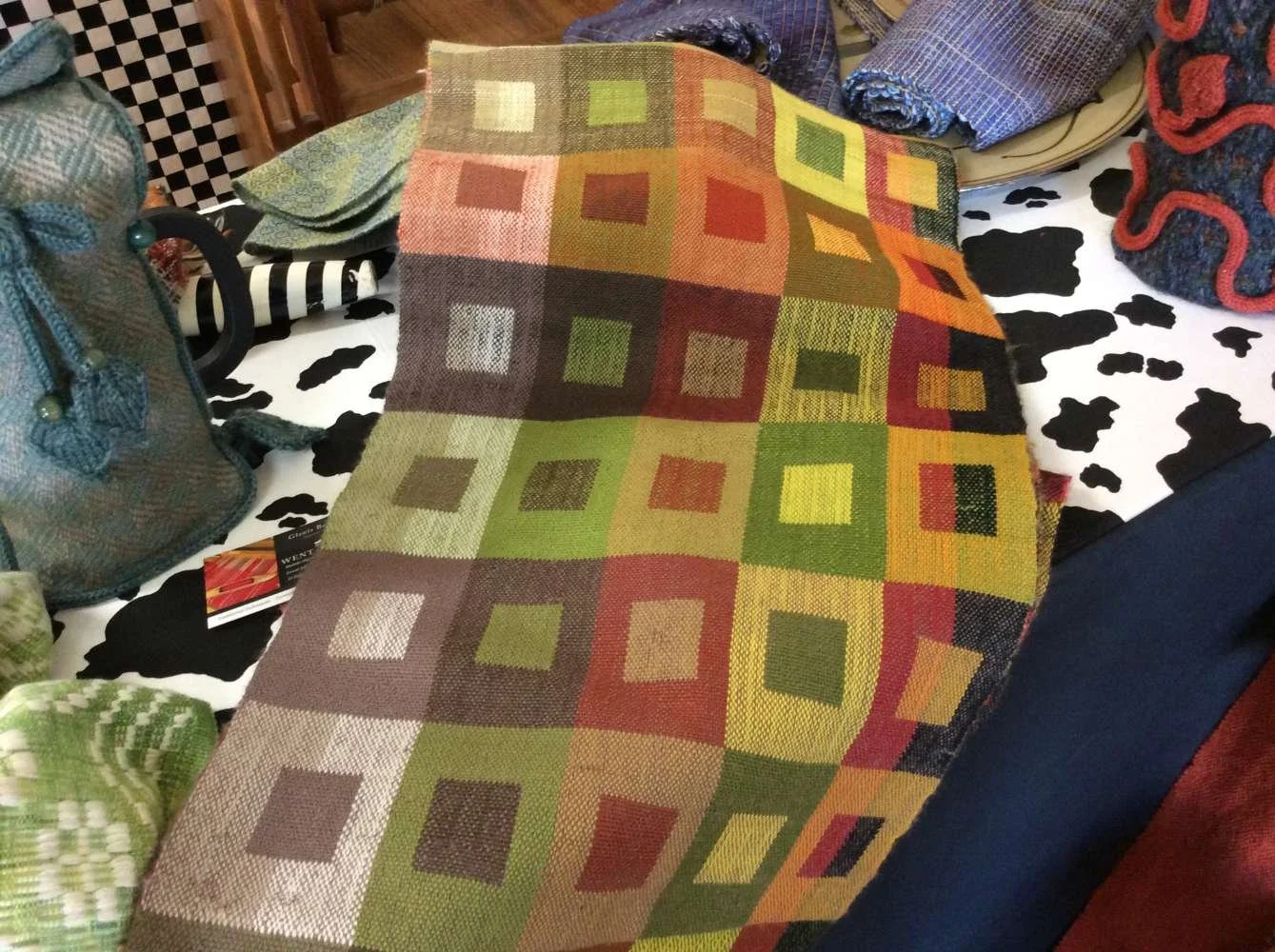 Colorful woven textile with a pattern of squares in various shades of green, red, and yellow, draped over a table with other fabric items visible around it.