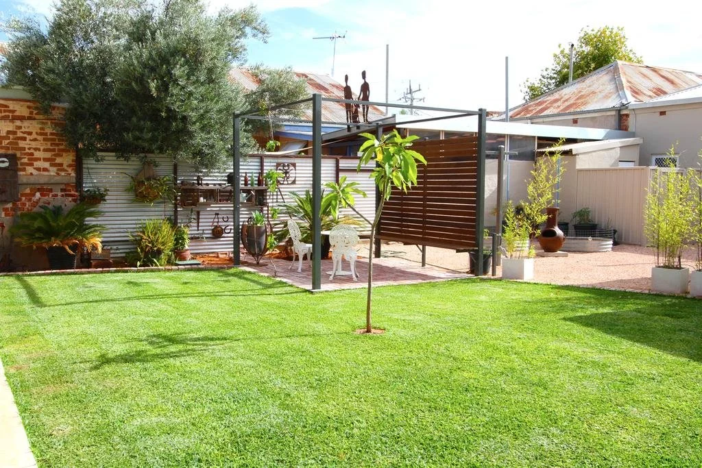 A backyard with well-maintained green grass, potted plants, a small tree, and a shaded patio area with garden furniture and decorative objects. Rustic buildings and a large tree are in the background.
