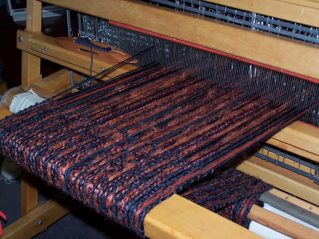 A weaving loom with a partially woven, multicolored fabric in shades of dark red, black, and brown.