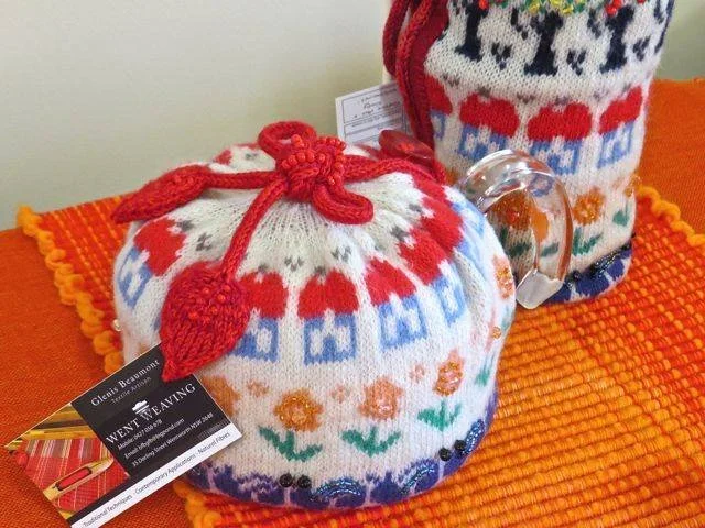 Knitted fabric bag with a red drawstring and pom-pom, decorated with colorful floral and pattern designs, placed on an orange textured surface.