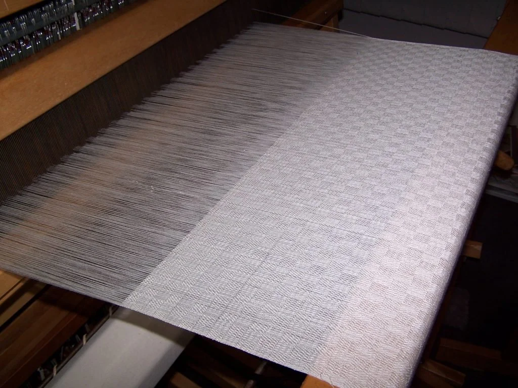 A weaving loom with a partially woven fabric in progress, showing the transition from dark colored threads to light colored threads.