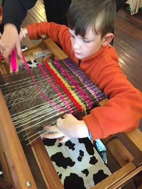 Child weaving a colorful textile on a loom with a person helping.