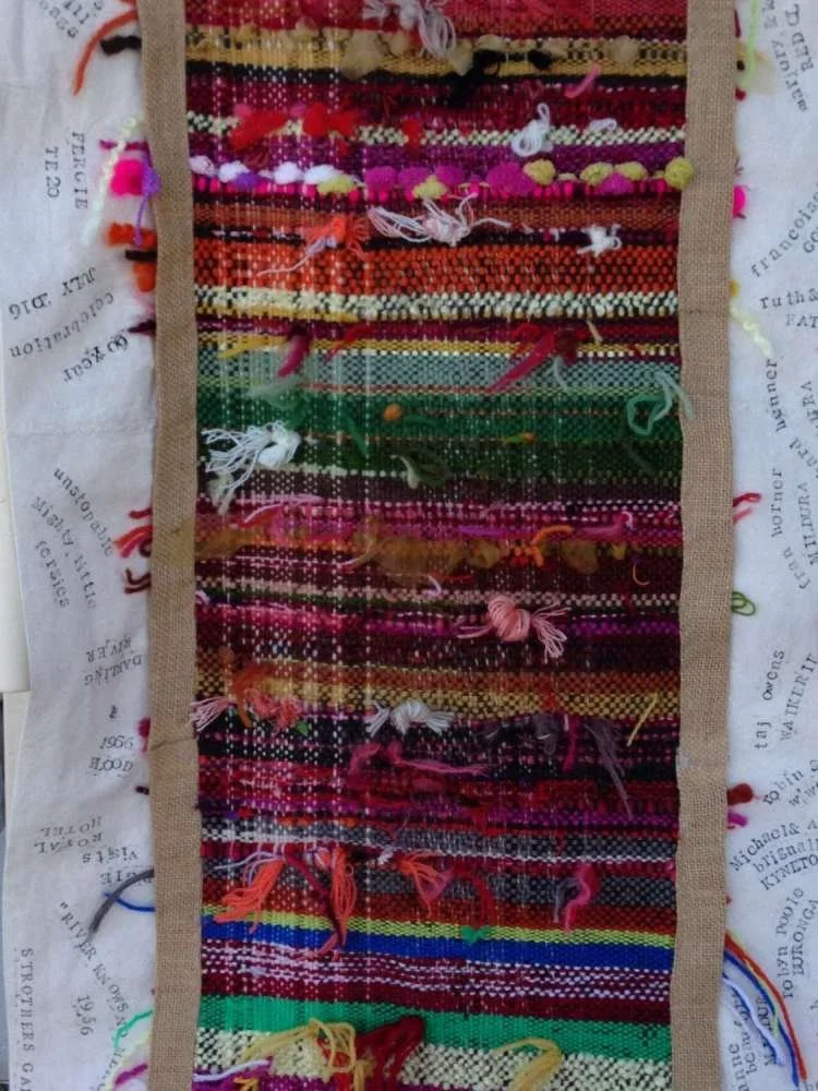 Colorful woven textile with frayed edges and scattered loose threads, laid on a background of printed fabric with text.