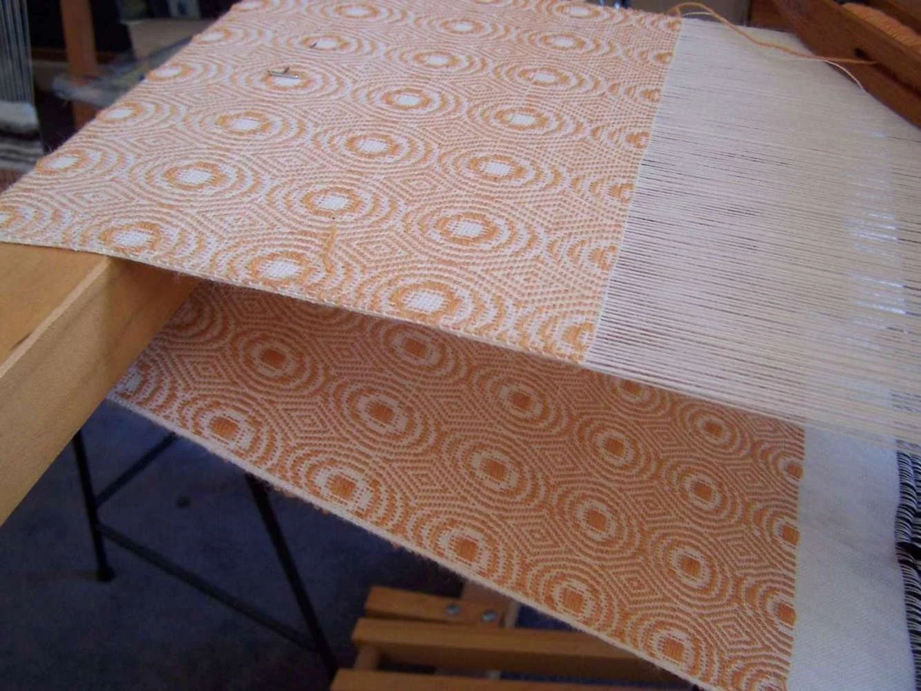 A textile loom with partially woven fabric featuring an orange and white geometric dot pattern.