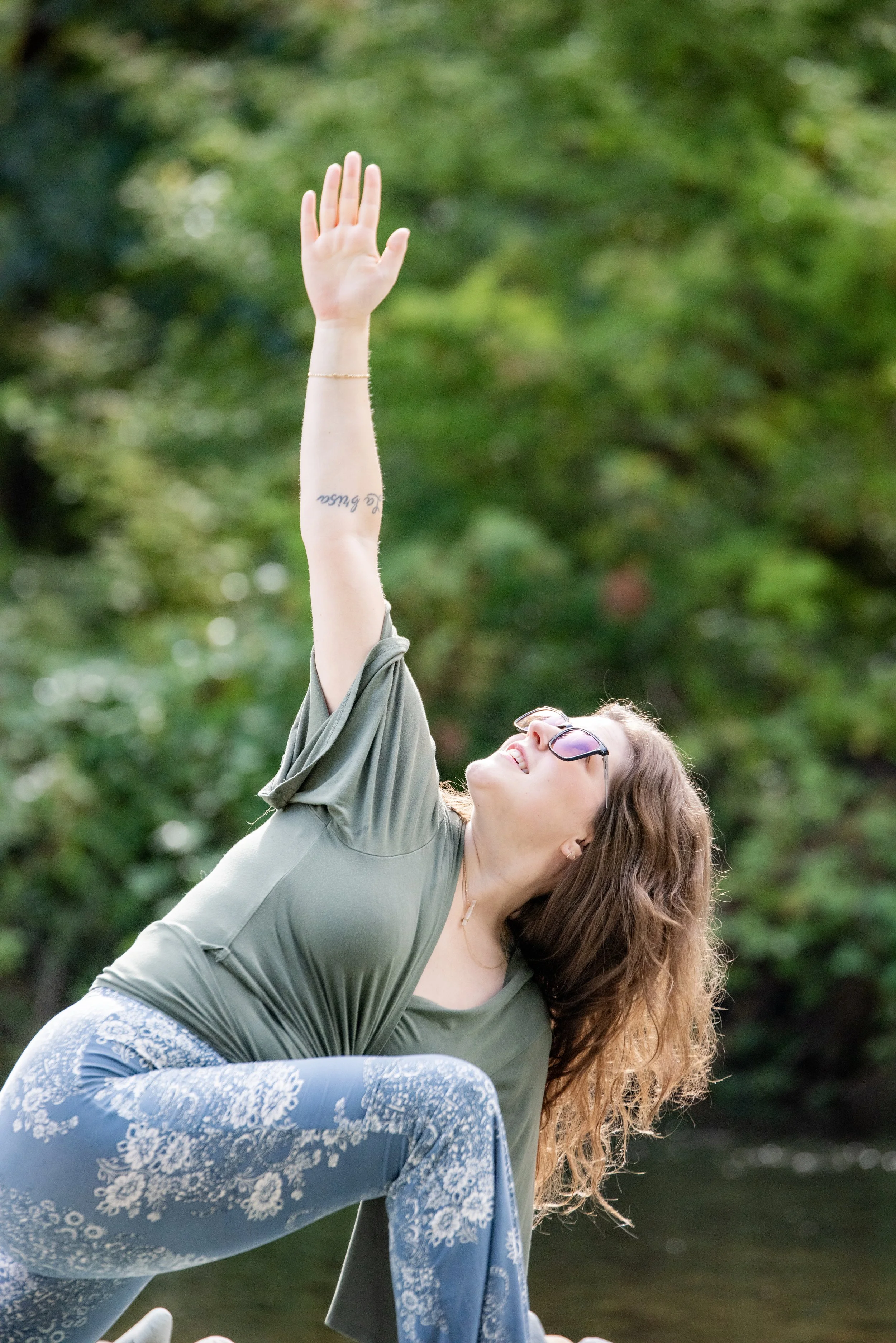 A woman with long, brown hair in a twisting low lunge yoga pose. Background includes nature scenery like trees and river.