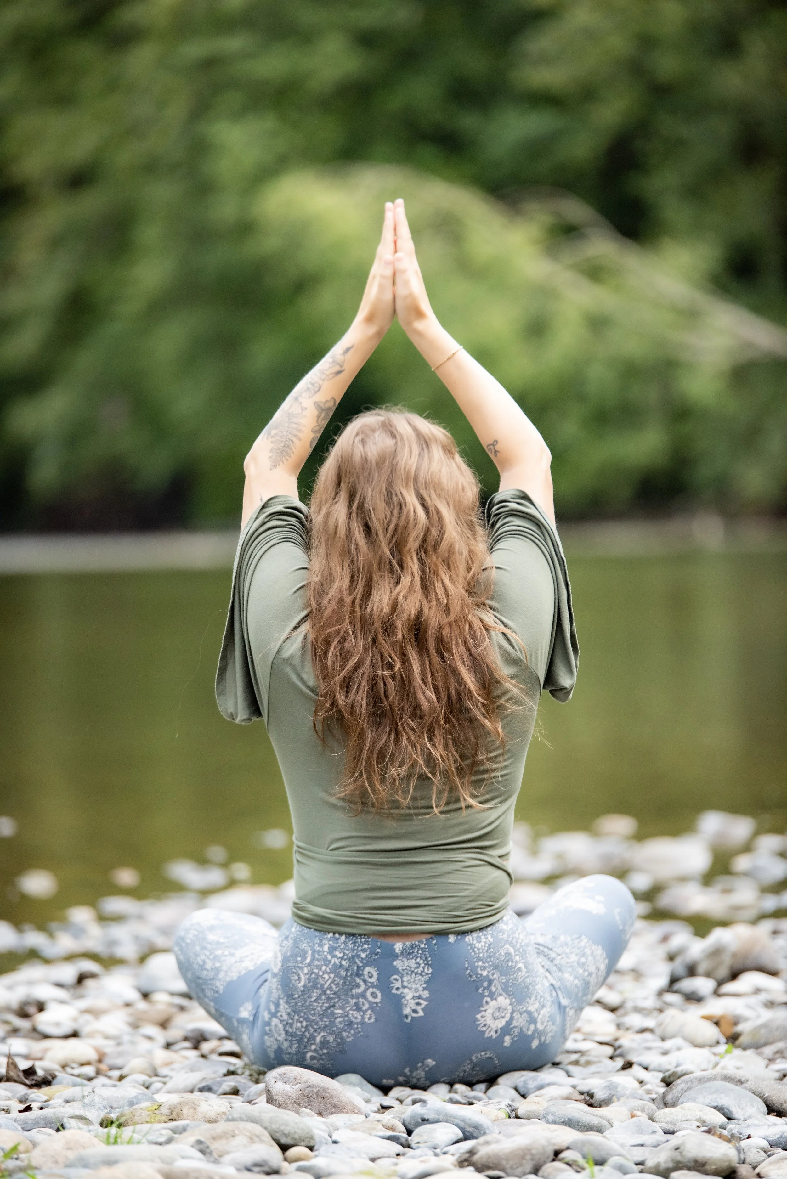 Behind view of woman seated cross-legged on rocks with arms touching overhead. Backdrop of nature scenery including trees and river.