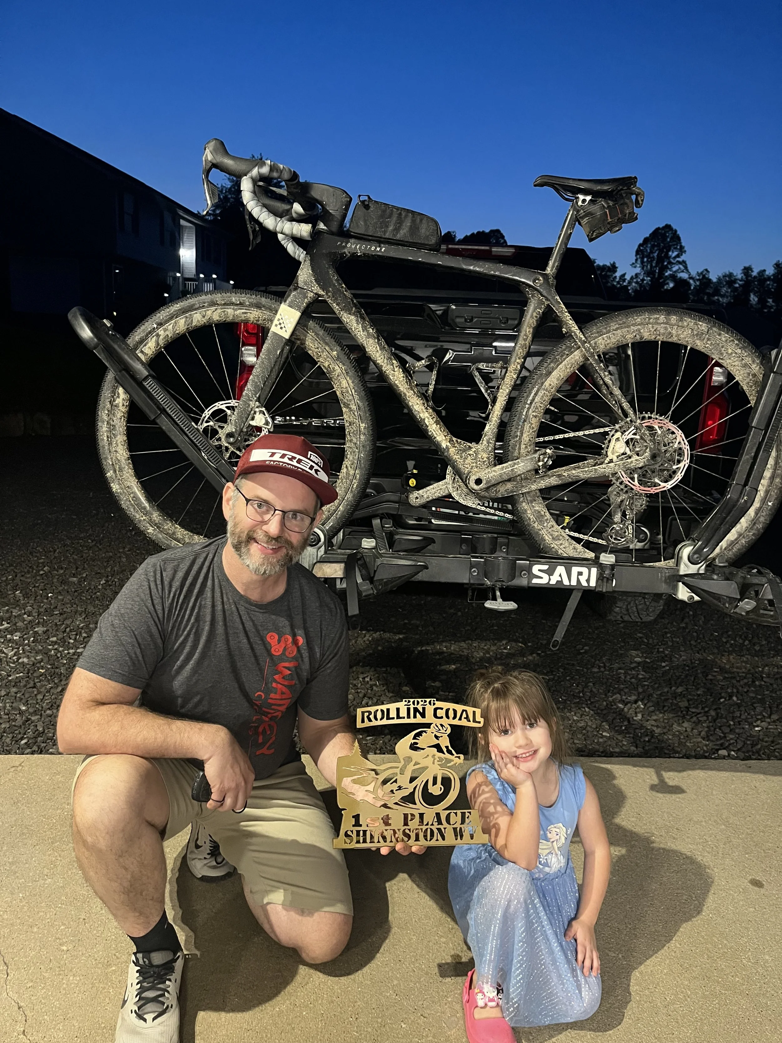 A man and a young girl kneeling on the sidewalk, holding a 1st place trophy for a mountain biking competition in front of a black pickup truck with a mountain bike on its roof. The man is wearing a gray shirt, shorts, and a red cap, while the girl is wearing a blue dress and pink shoes. The scene is outdoors during nighttime, with trees and buildings in the background.