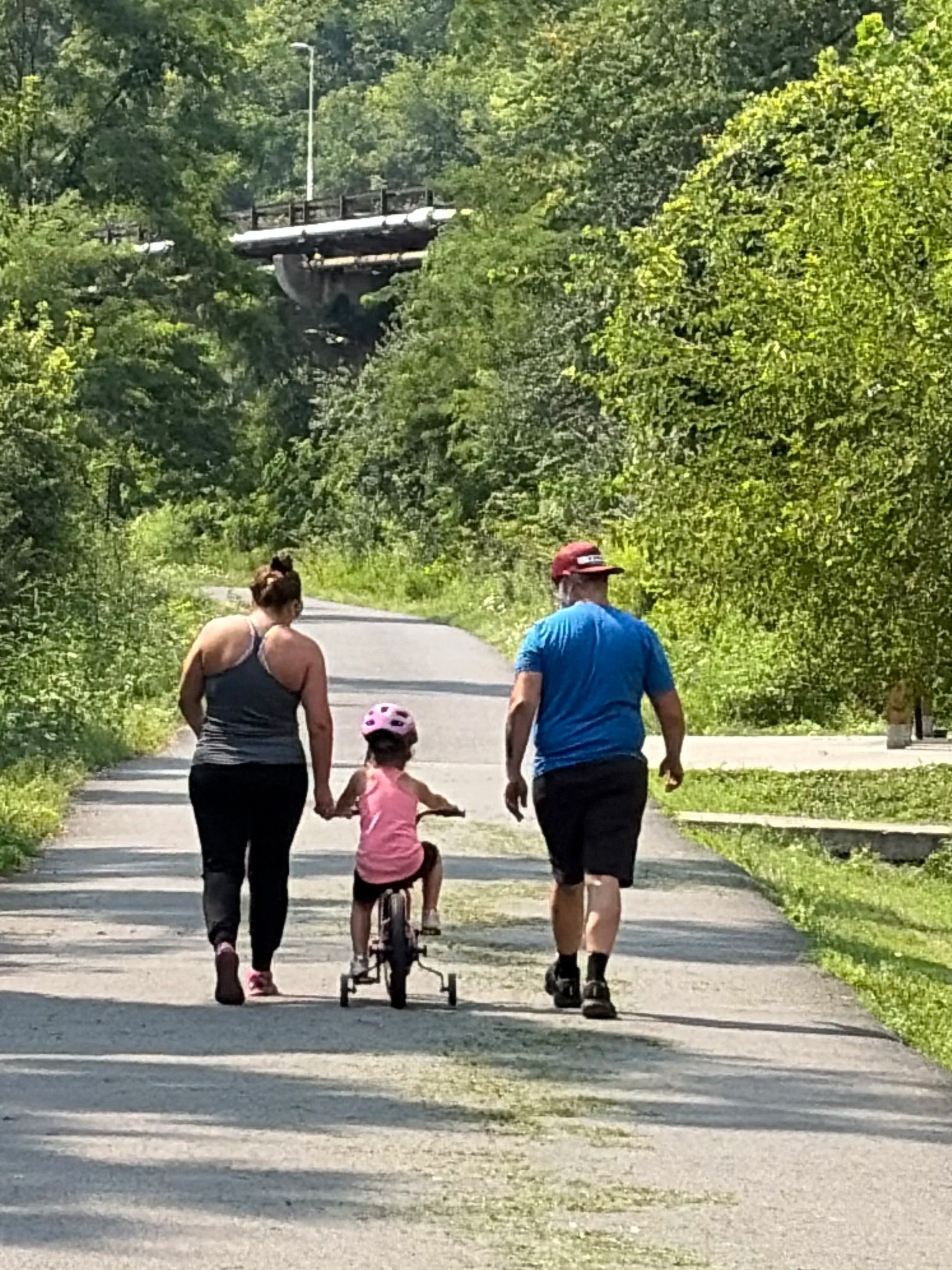 A family of four, including two adults and a child on a bike, walks on a paved trail in a green, wooded area on a sunny day.
