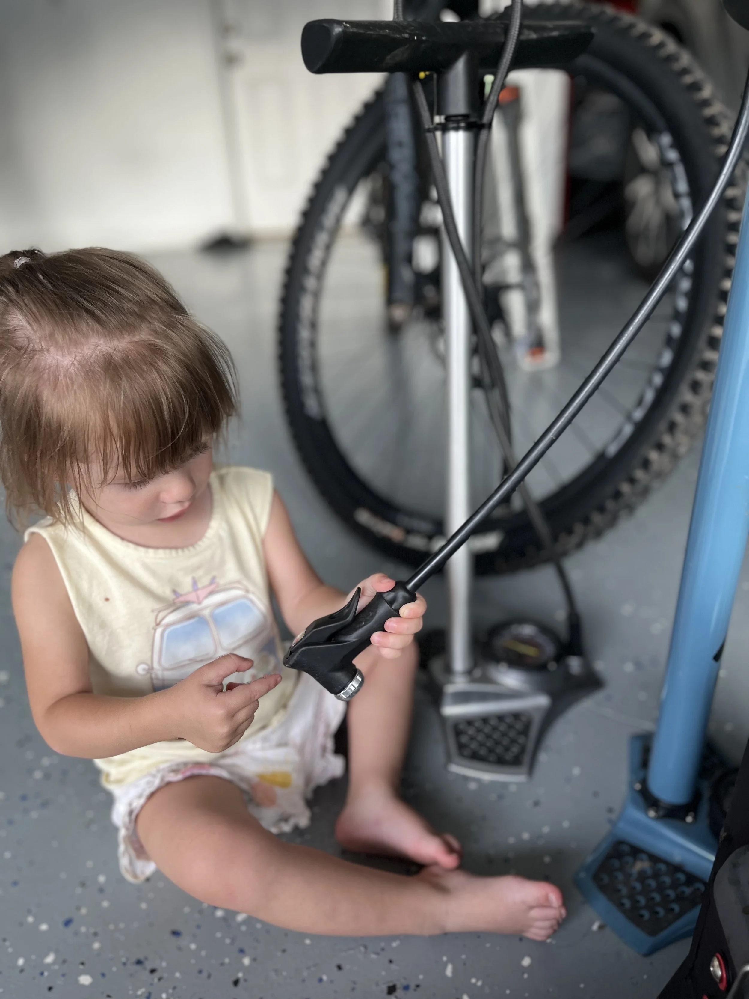 A young girl with brown hair, wearing a sleeveless light yellow shirt with a cartoon design, is sitting on the floor. She is holding a remote control, and a black bike with thick tires is visible in the background. The floor is gray with small speckled patterns.