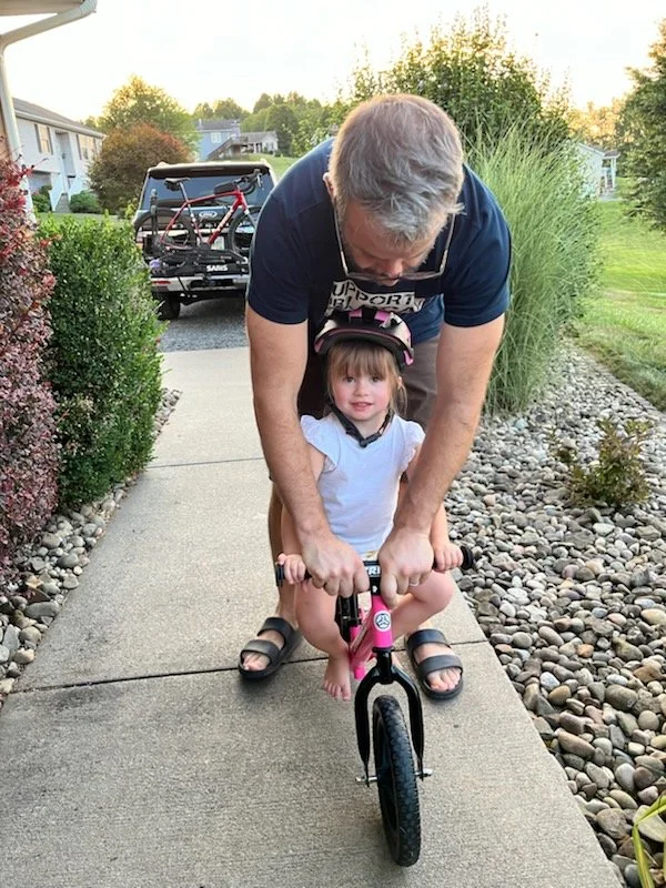 A man helping a young girl learn to ride a pink balance bike on a sidewalk in a residential neighborhood during sunset.