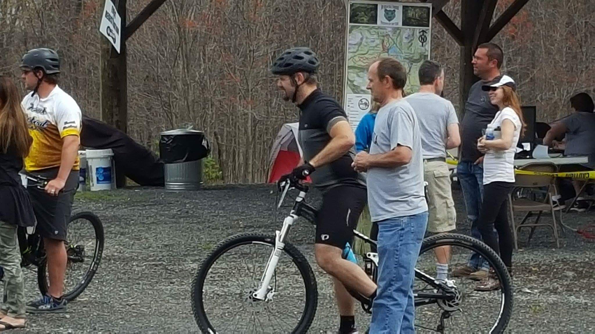 Group of people gathered at an outdoor event, some with bicycles and wearing helmets. A man and woman are engaged in conversation, and there are informational signs in the background.