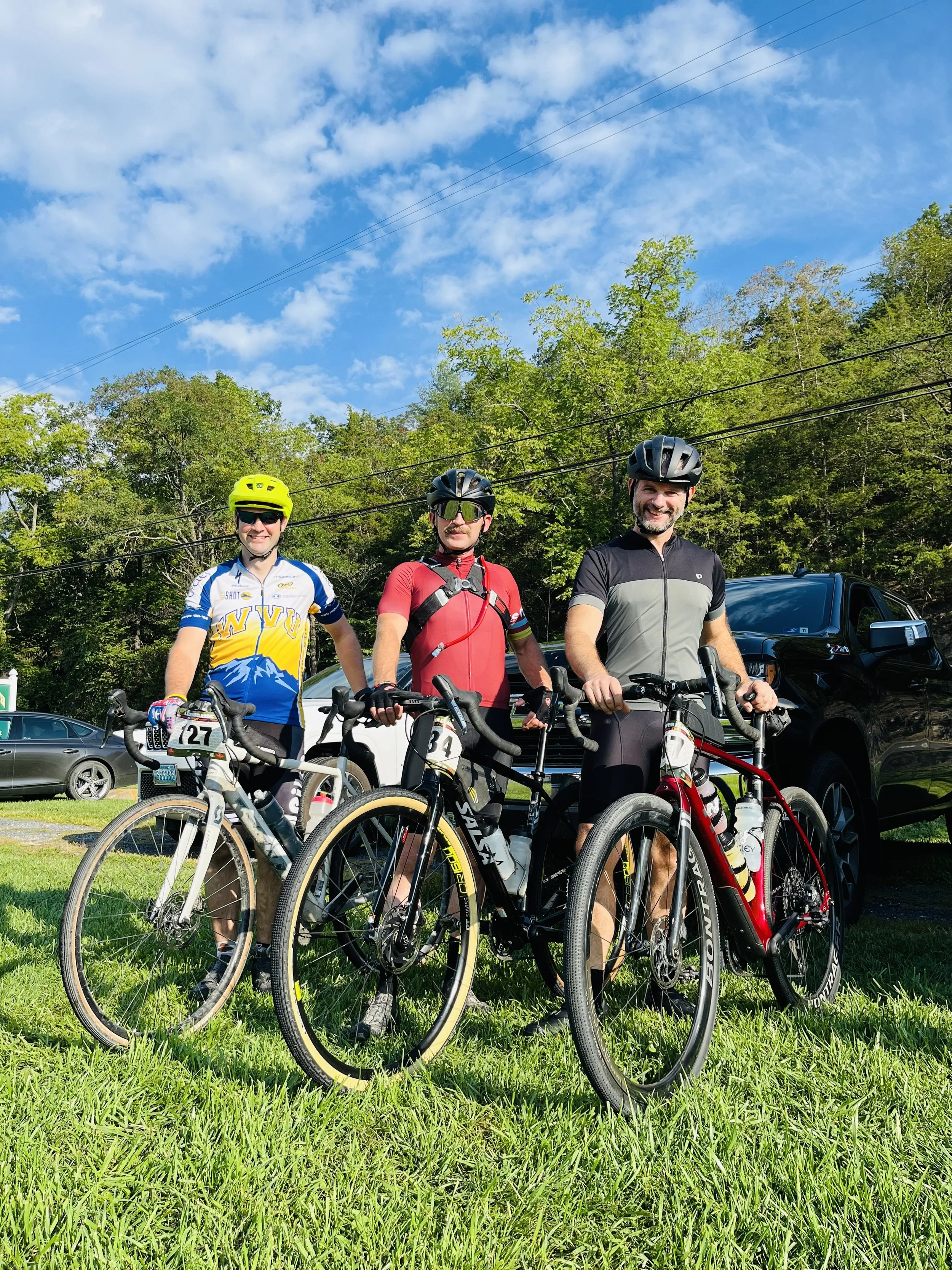 Three men in cycling gear and helmets standing with their bikes on a grassy area with trees, parked cars, and a blue sky with some clouds in the background.