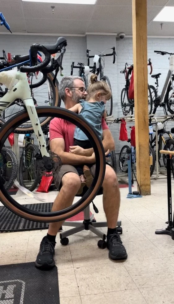 A person with glasses and a beard sitting on a stool in a bike shop, holding a young girl on their lap. The girl has her arms around the person's neck. Bicycles are visible in the background.