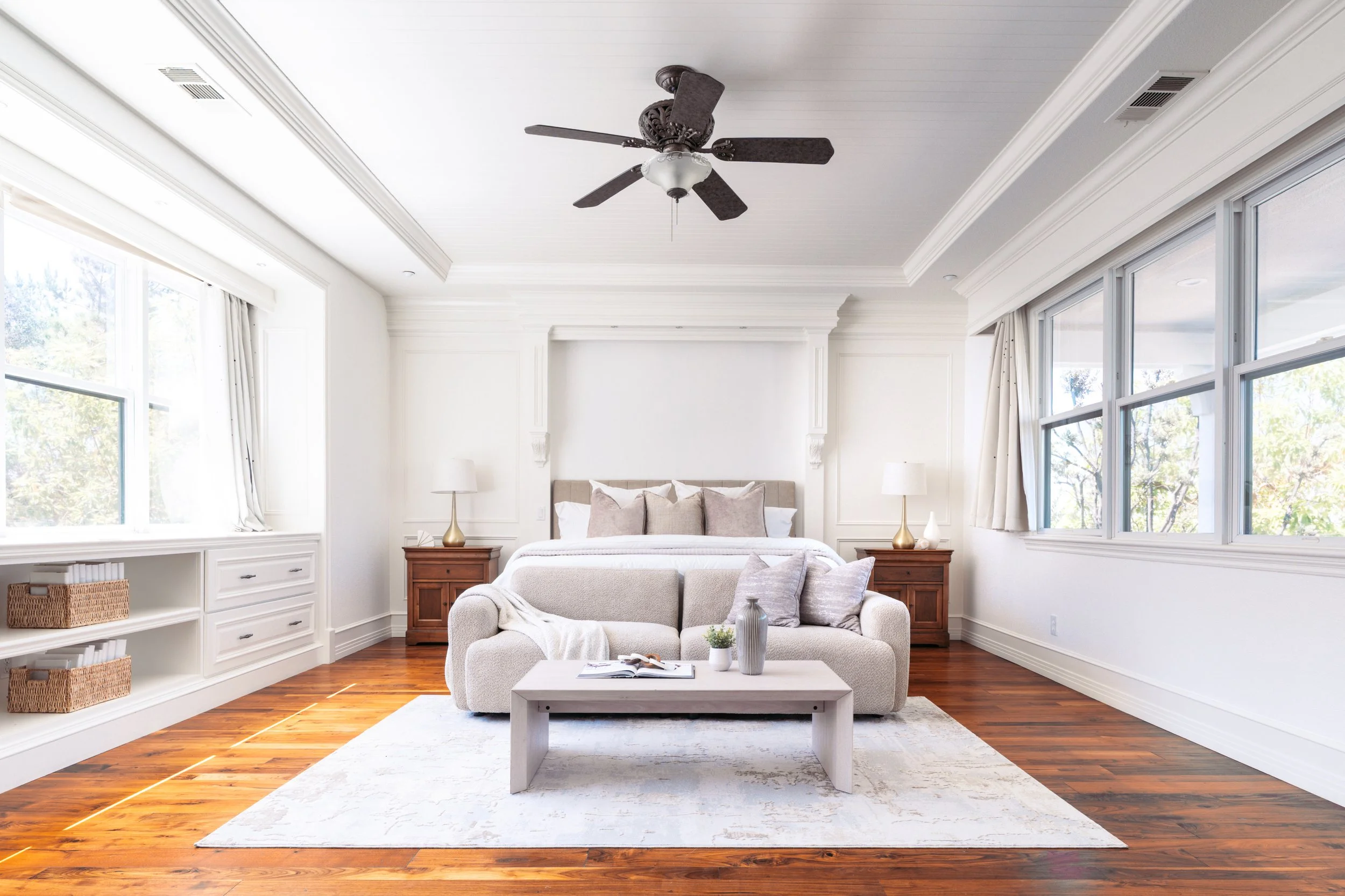 Bright living room with large windows, white walls, a beige sofa with pillows, brown hardwood floors, wooden side tables with lamps, a white coffee table with a book and vases, and a ceiling fan.
