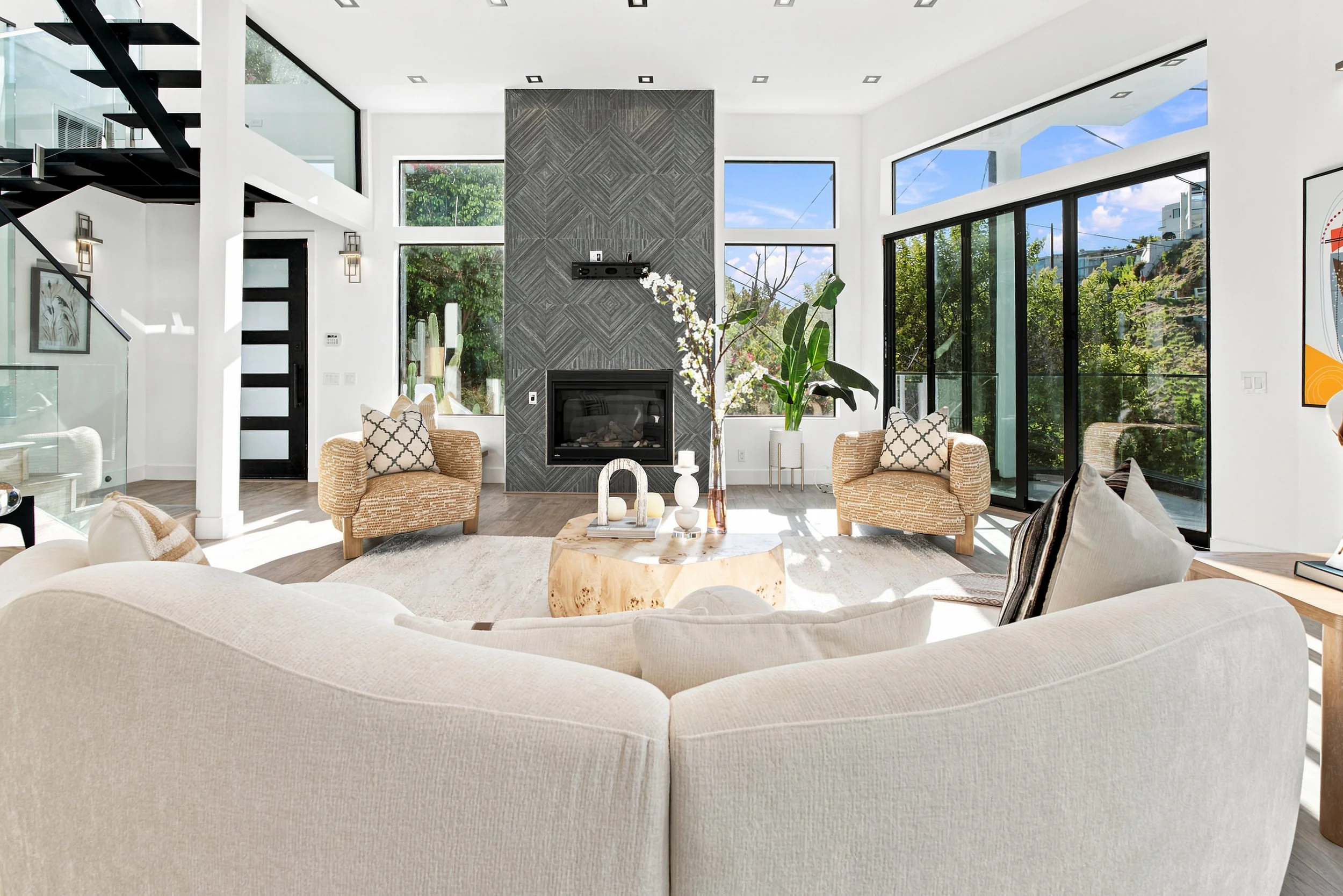A spacious living room with a large cream-colored sectional sofa, a circular wooden coffee table, and a gray stone fireplace. The room has floor-to-ceiling windows showing a lush outdoor view, and modern decor with plants and artwork.