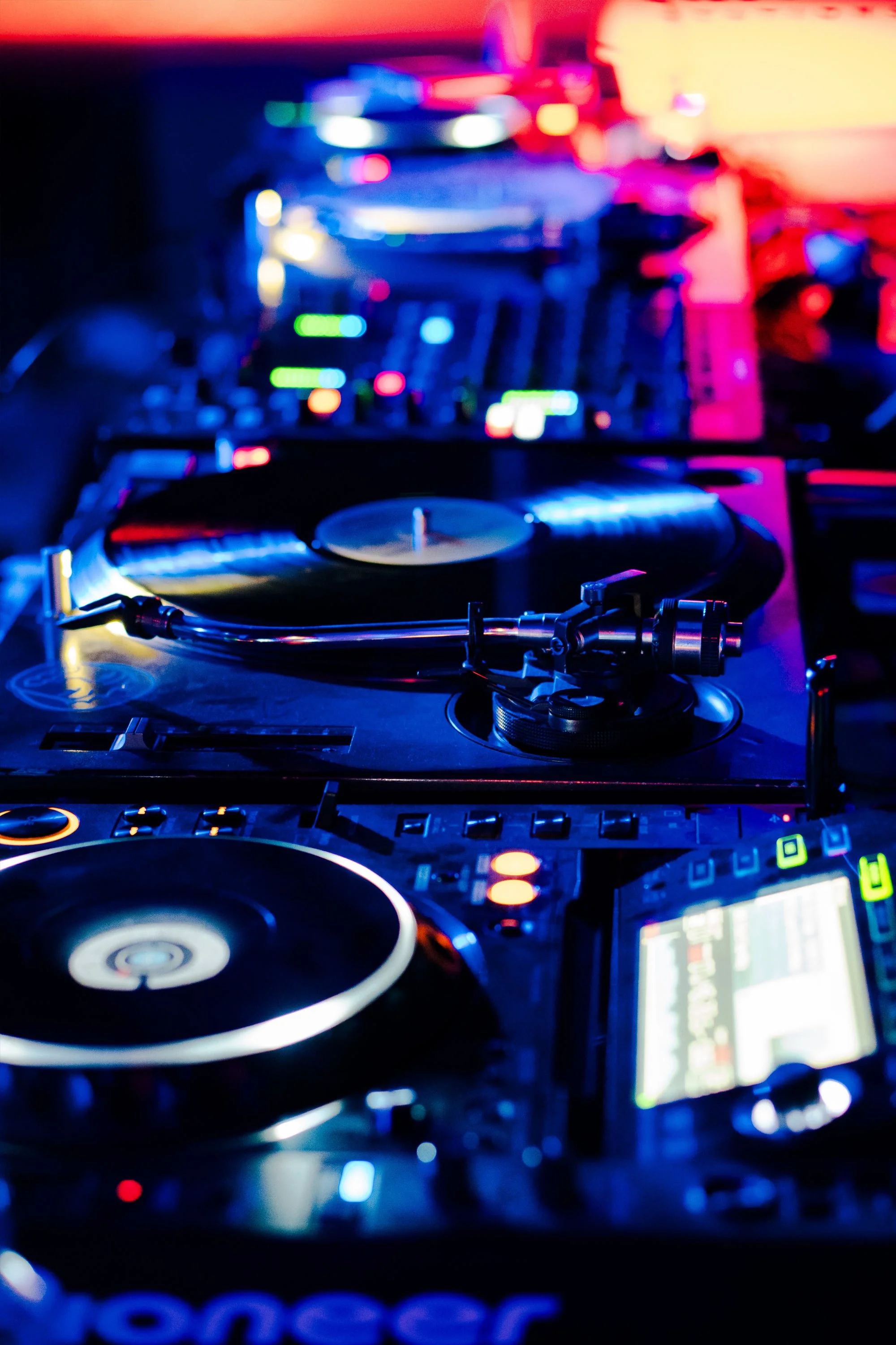 Mesa de mezcla de DJ con controles, tocadiscos, consola y pantallas, iluminada con luces de colores brillantes en un ambiente de fiesta o club.