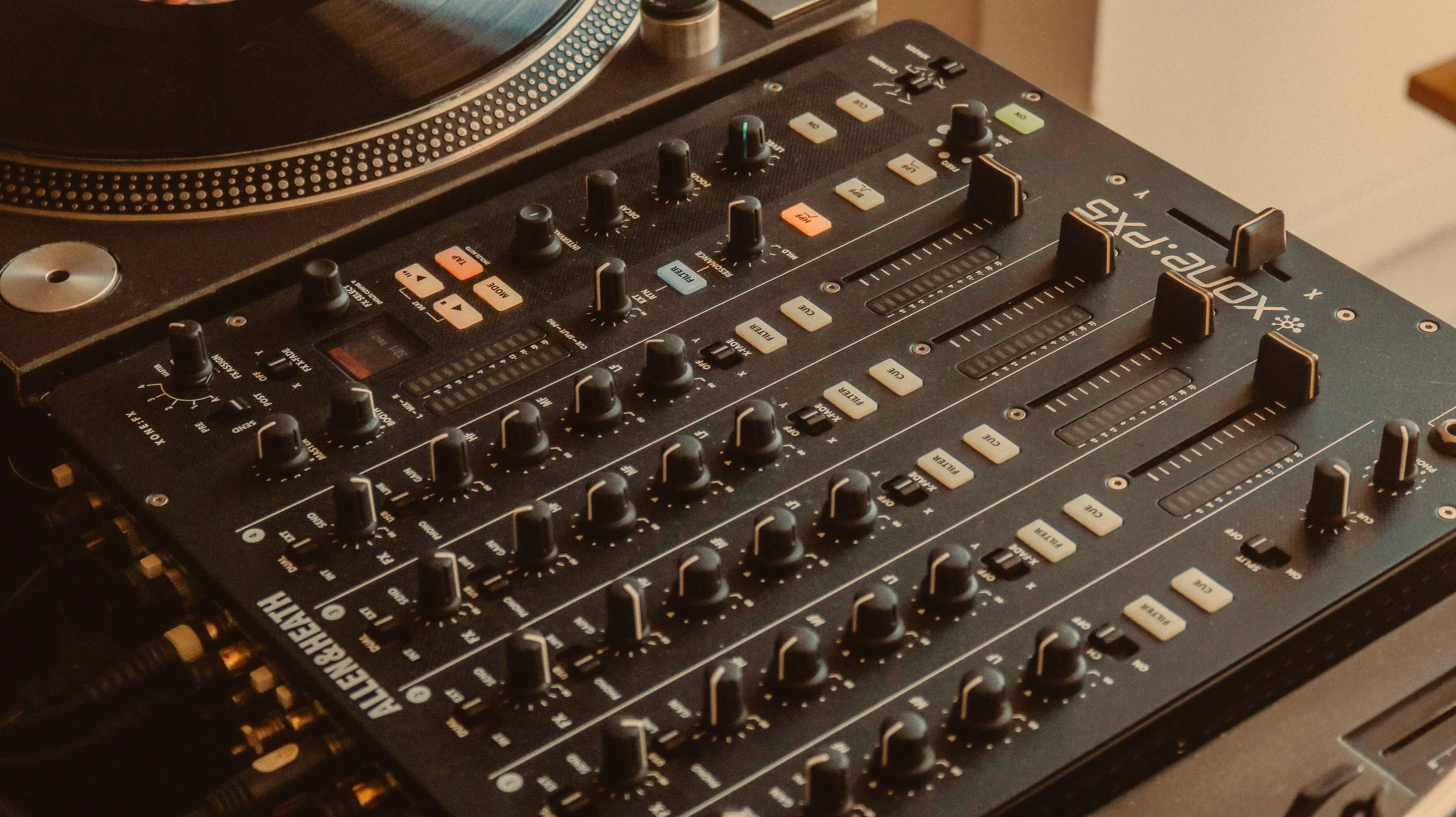 Una mixer consola de mezclas de DJ  ALLEN HEATH xone px5 con múltiples controles y botones, con un plato tocadiscos dj technics sl1200 MKII giratorio parcialmente visible a la izquierda.