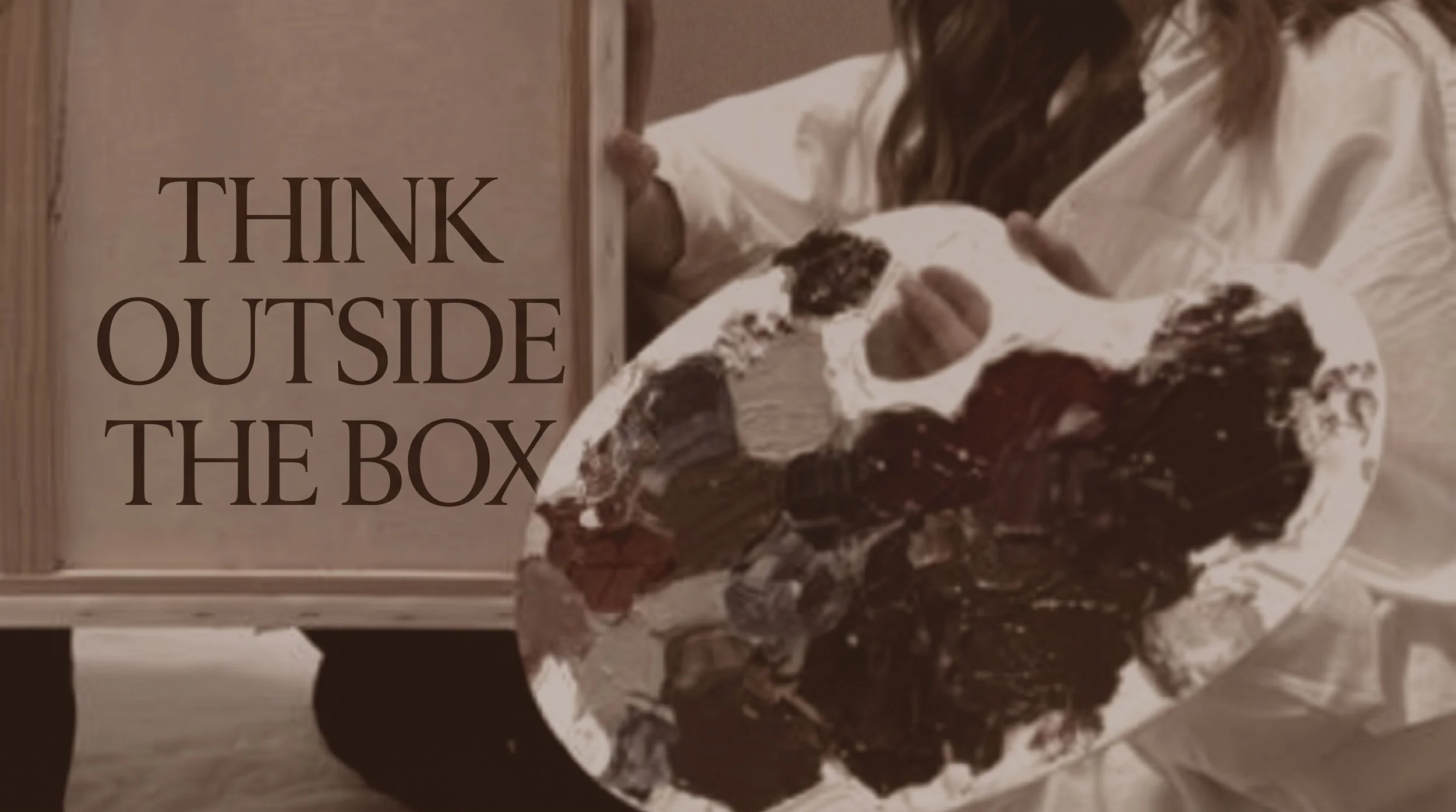 A person holding a white box filled with assorted chocolates and flowers, standing next to a wooden sign that reads "THINK OUTSIDE THE BOX".