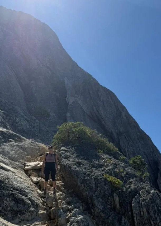 Hiker walking on a rocky mountain trail with a large mountain face in the background under a clear blue sky.