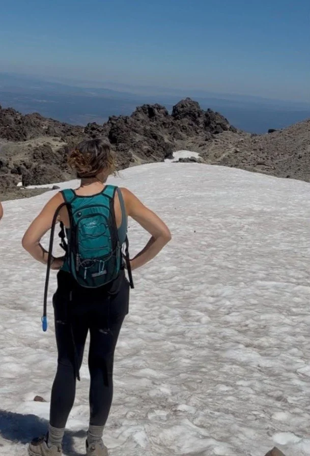 A woman with a backpack standing on a snowy mountain trail, looking at rocky terrain and distant landscape.