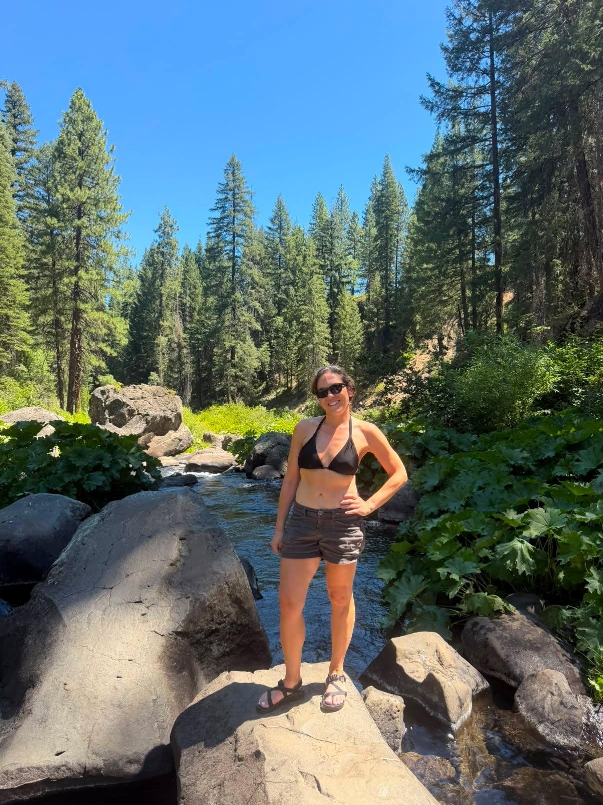 A woman in a black bikini top, shorts, and sandals standing on a large rock in a forested creek, smiling with sunglasses, surrounded by green trees and rocks under a clear blue sky.