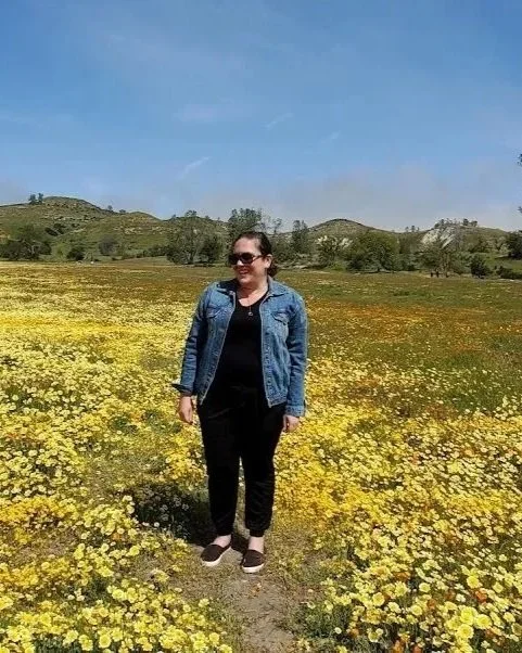 Woman wearing sunglasses, a denim jacket, and black pants standing in a field of yellow flowers with hills and trees in the background under a blue sky.