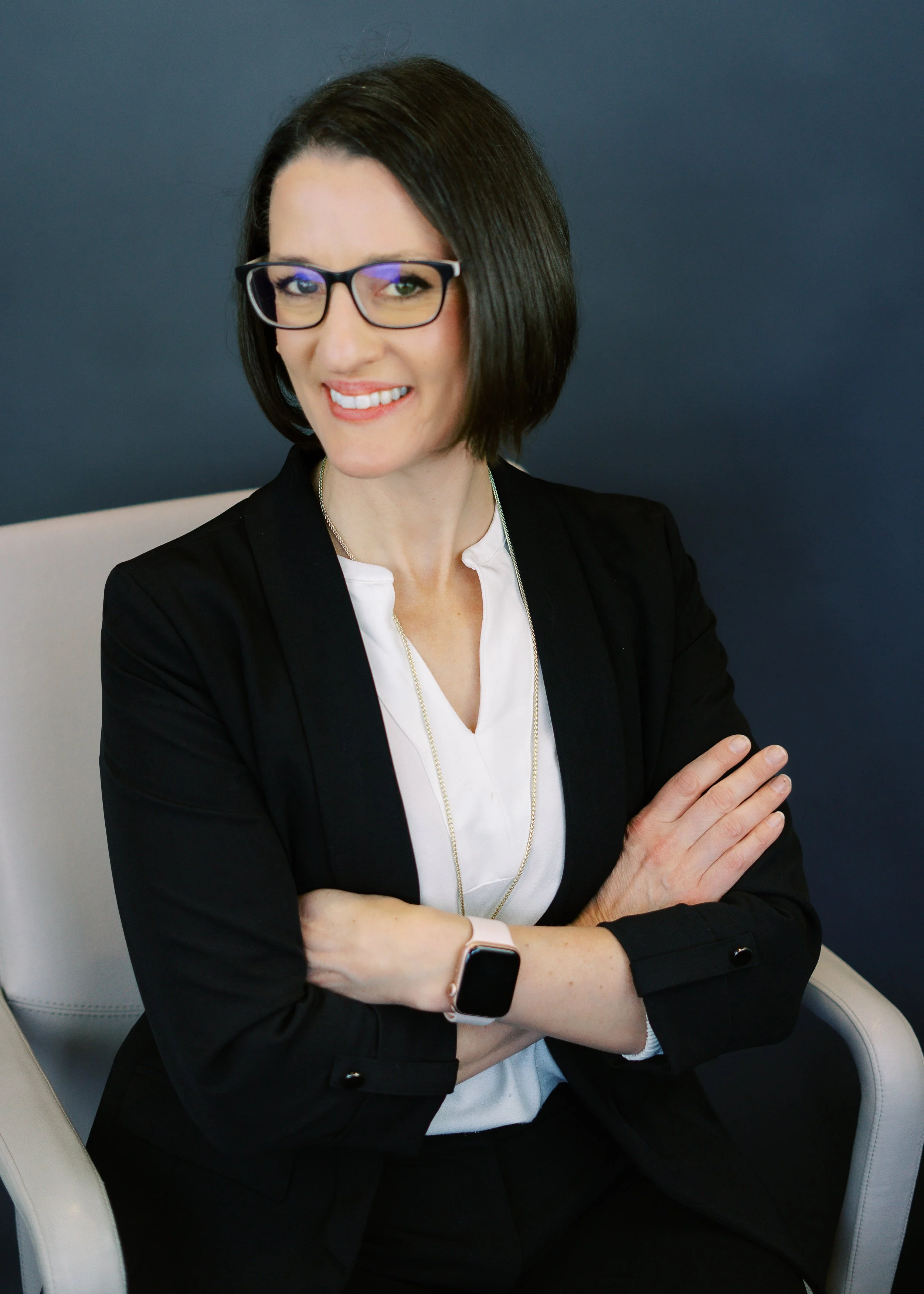 A woman with short dark hair, wearing glasses, a black blazer, and a white blouse, sitting with crossed arms and smiling at the camera against a dark blue background.