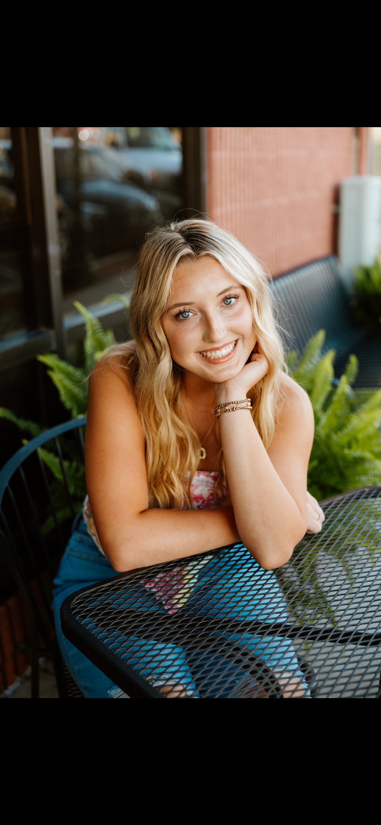 A young woman with long blonde wavy hair and blue eyes, smiling, sitting at an outdoor cafe table surrounded by green plants. She is resting her head on her hand and wearing a colorful top, denim shorts, and bracelets.