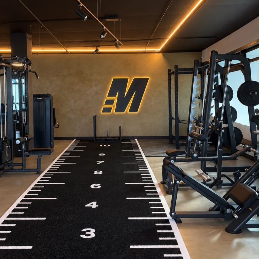 Indoor gym featuring workout machines, free weights, and a black turf with white yard and meter markings, illuminated by modern lighting, with a large illuminated logo on the beige wall at the back.