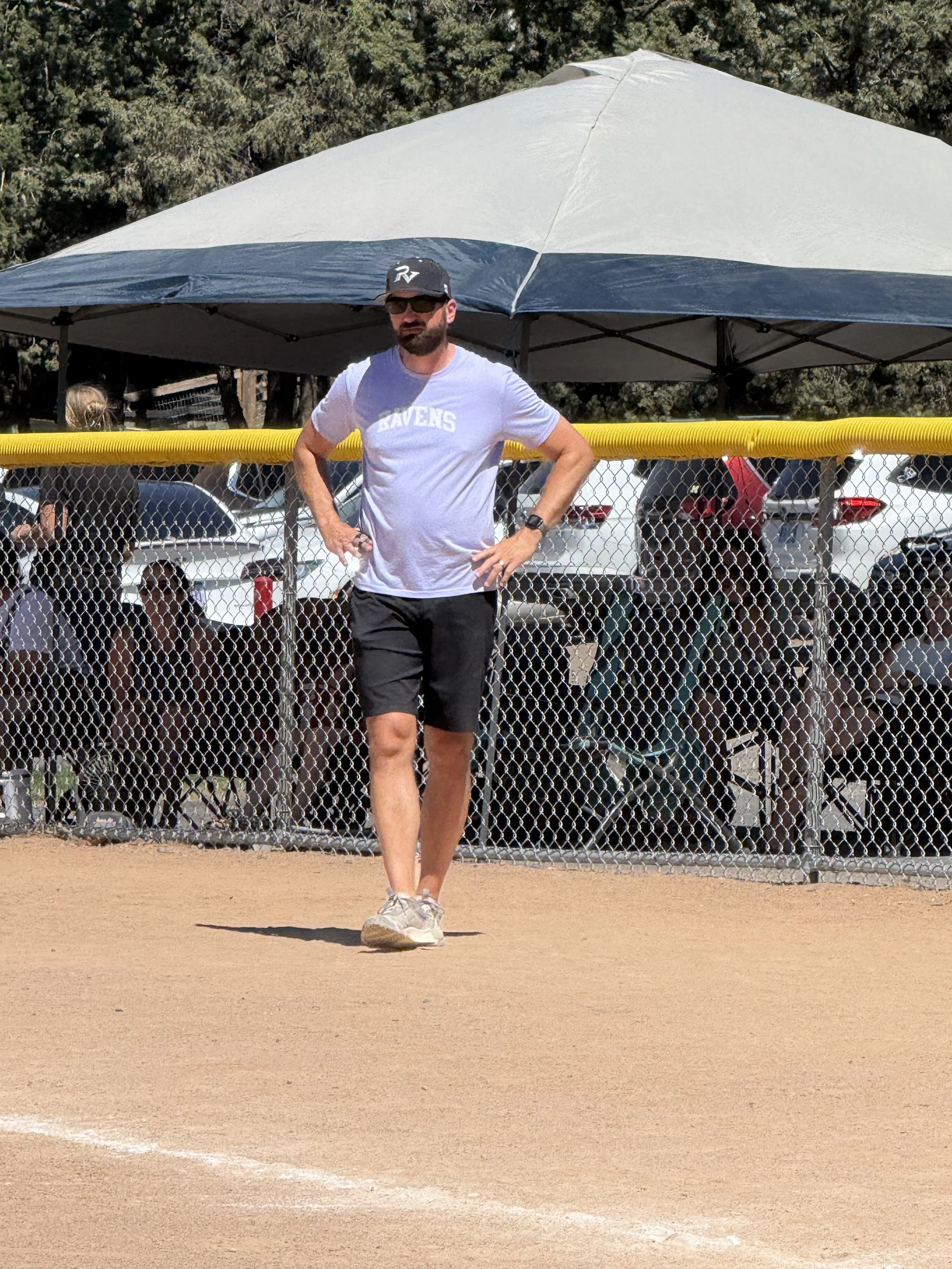 Man standing on a baseball or softball field's dirt infield, wearing sunglasses, a black cap, a white T-shirt with the word 'RAVENS,' black shorts, gray sneakers, and a watch, with a yellow safety fence and a tent behind him, and spectators in the background.