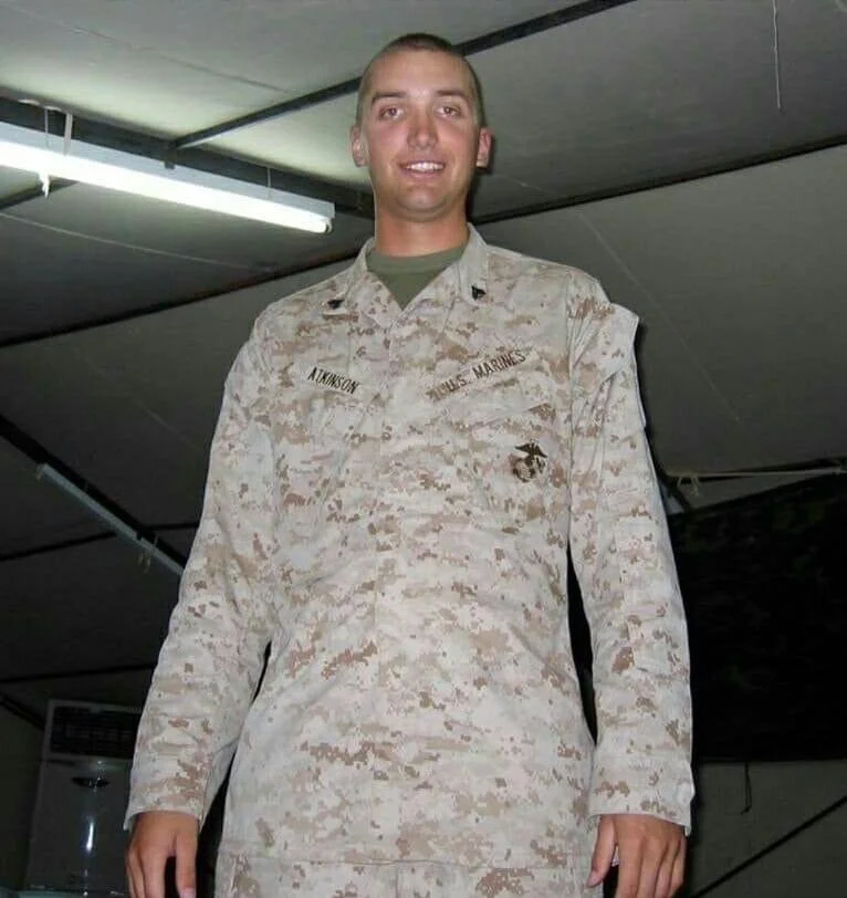 A young man in a camouflage Marine uniform standing indoors, smiling at the camera.