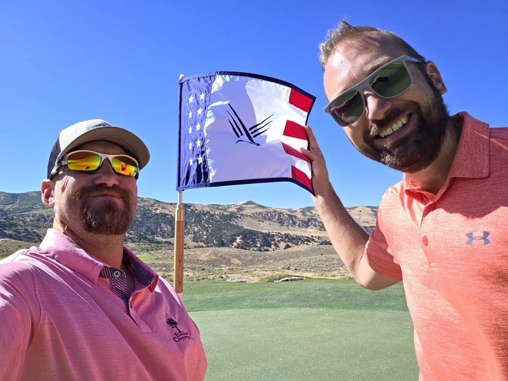 Two men on a golf course, smiling at the camera, with a flag in the background. The man on the left is wearing sunglasses, a pink polo shirt, and a cap. The man on the right is wearing sunglasses and a coral polo shirt.