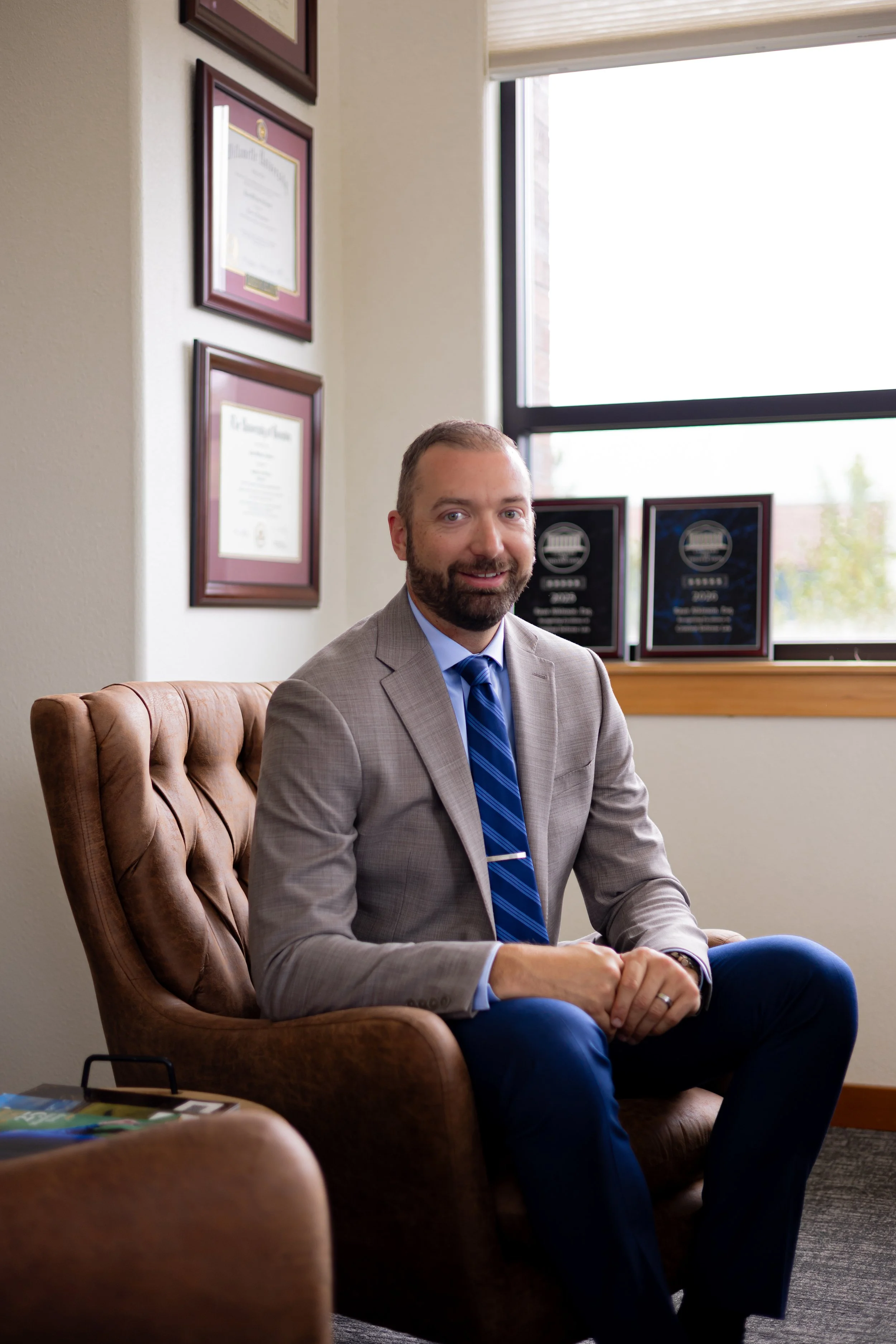 A man in a gray suit and blue tie sitting on a tan leather armchair in an office. Behind him are framed certificates on the wall and a large window with a view outside. The man is smiling and looking at the camera.
