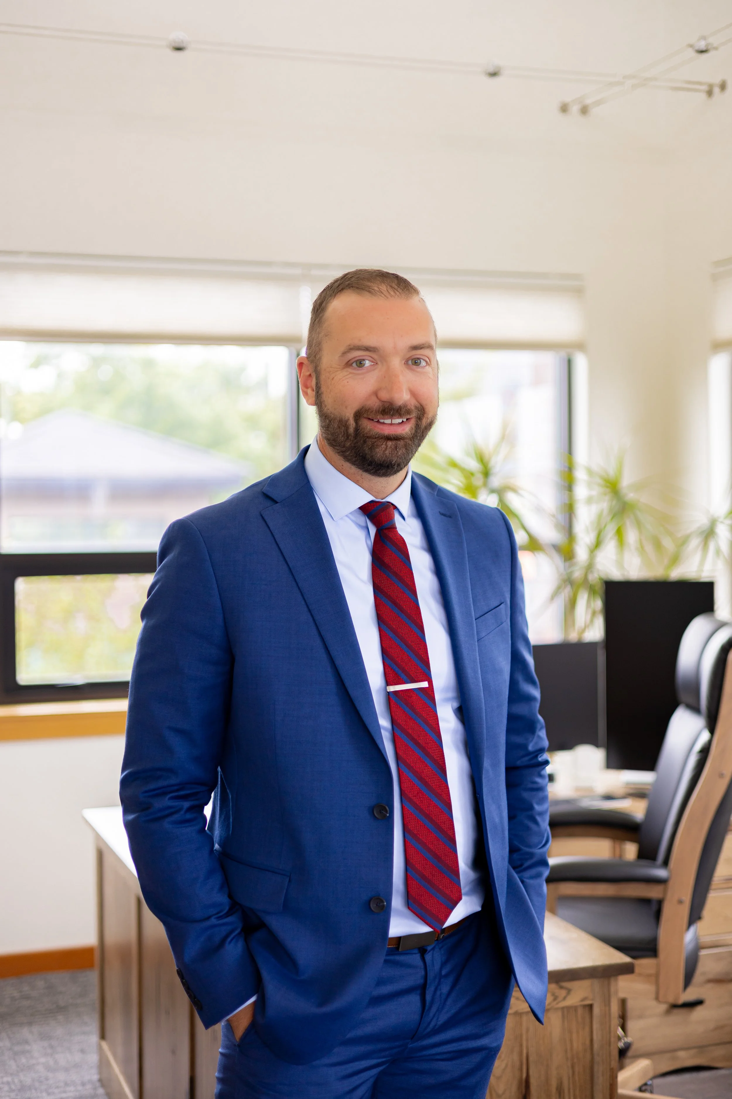 A man with a beard smiling and wearing a blue suit, white shirt, and red striped tie, standing in an office with large windows and computer desks.
