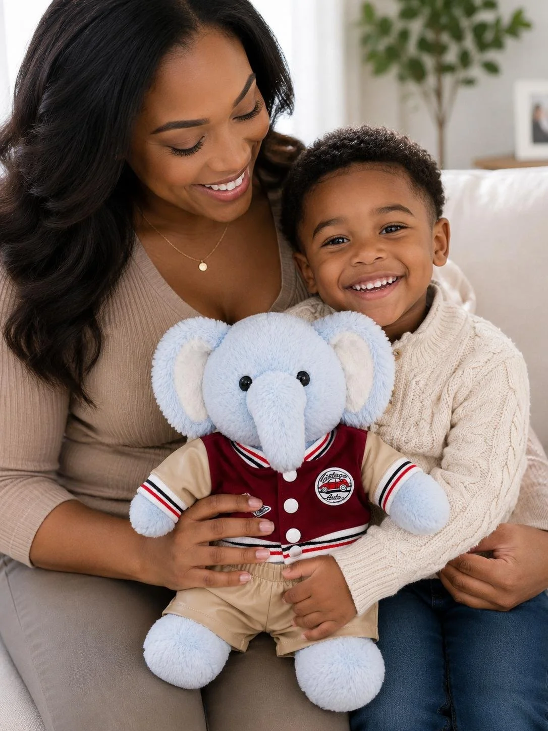 A smiling young woman and a young boy sitting on a couch, holding a light blue stuffed elephant toy dressed in a red and beige jacket with an auto repair logo, in a cozy living room with a plant and a framed photo in the background.
