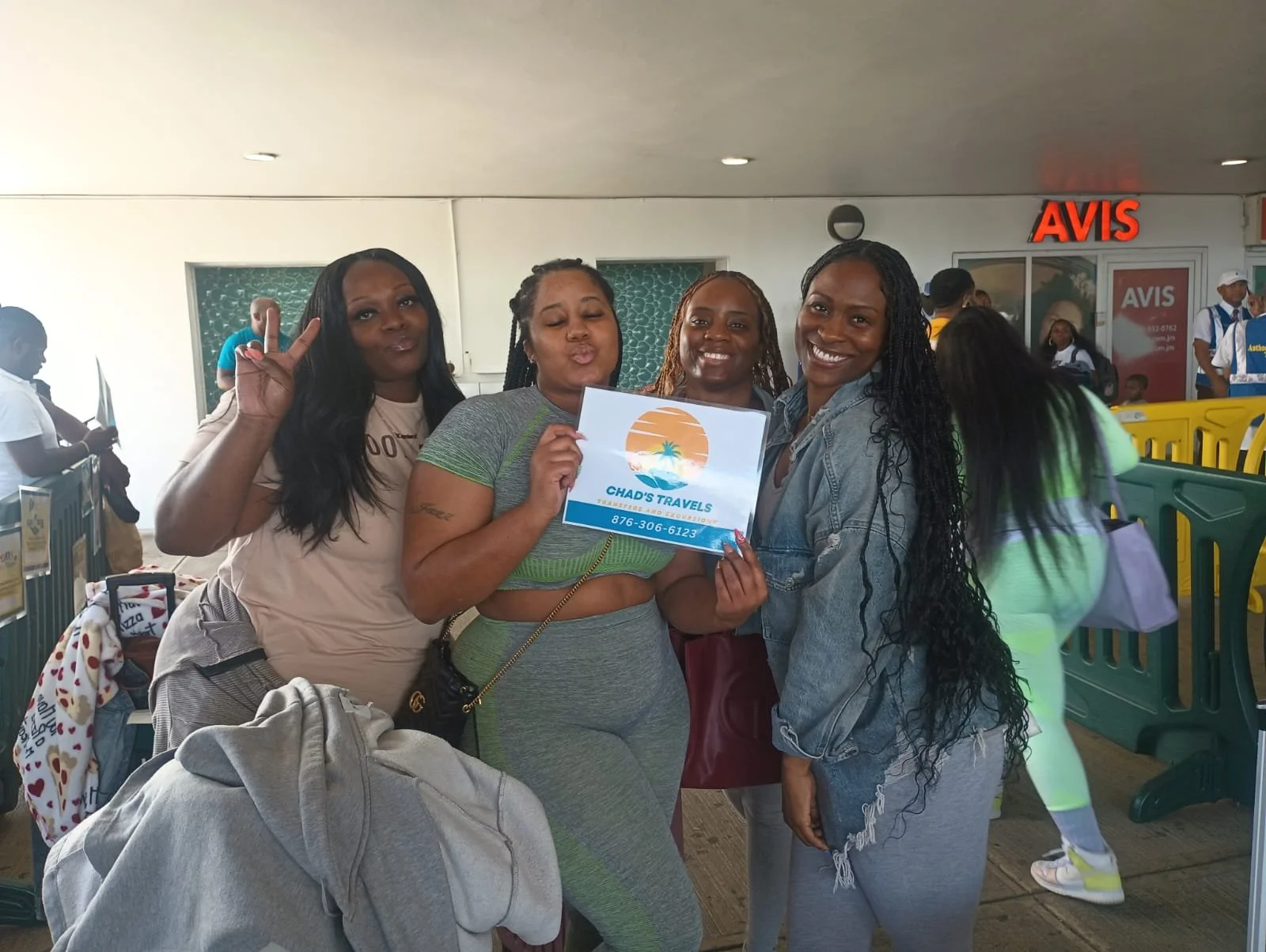 Four women at an airport, one holding a sign that reads 'Chad's Travels' with a sunset, palm trees, and ocean graphic. They are smiling and making peace signs.
