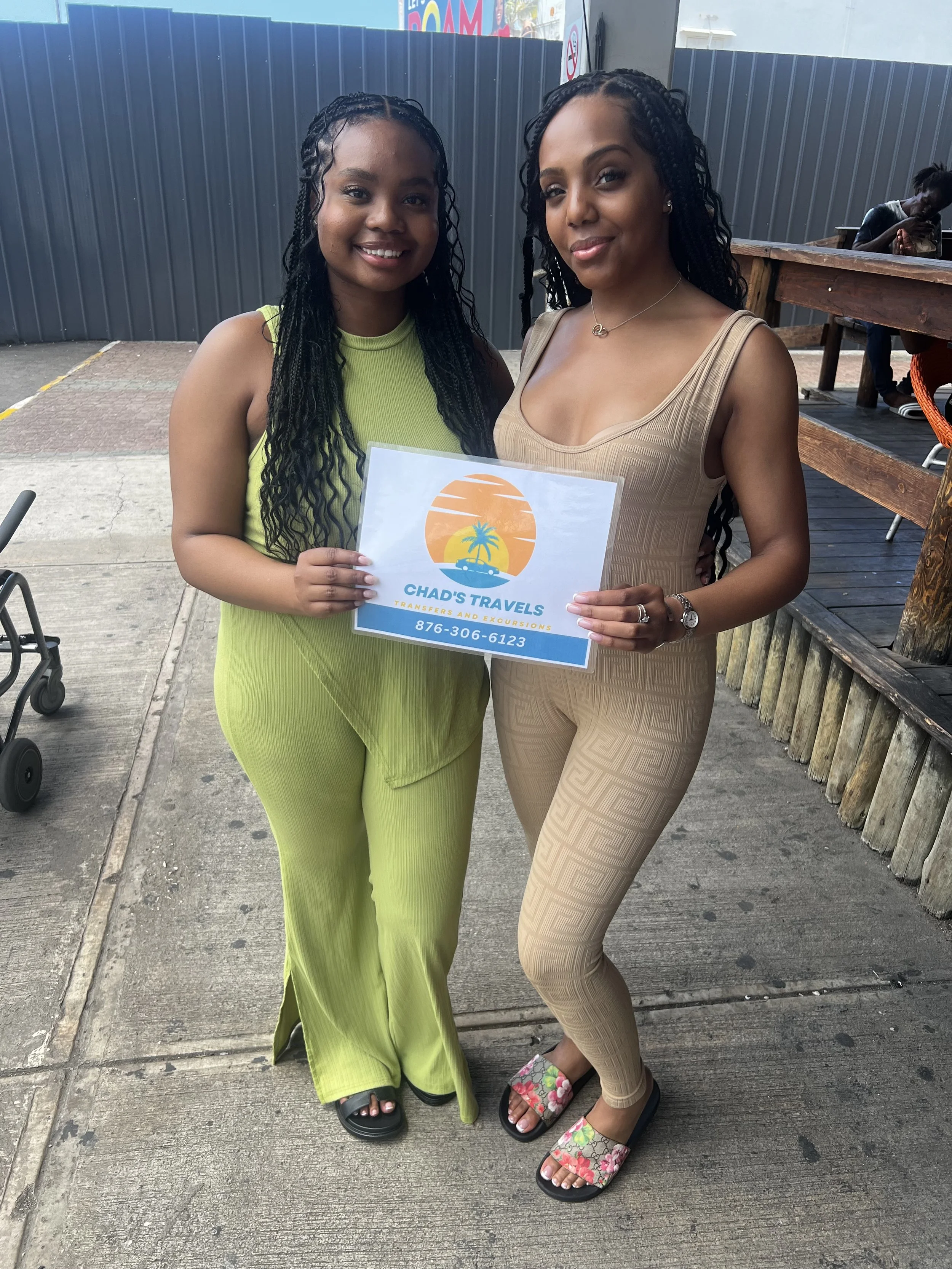 Two women posing outdoors, one in a bright green dress and the other in beige patterned outfit, holding a sign for Chad's Travels, with a background of a dark gray fence and wooden deck, one woman wearing floral slide sandals and the other black slides.