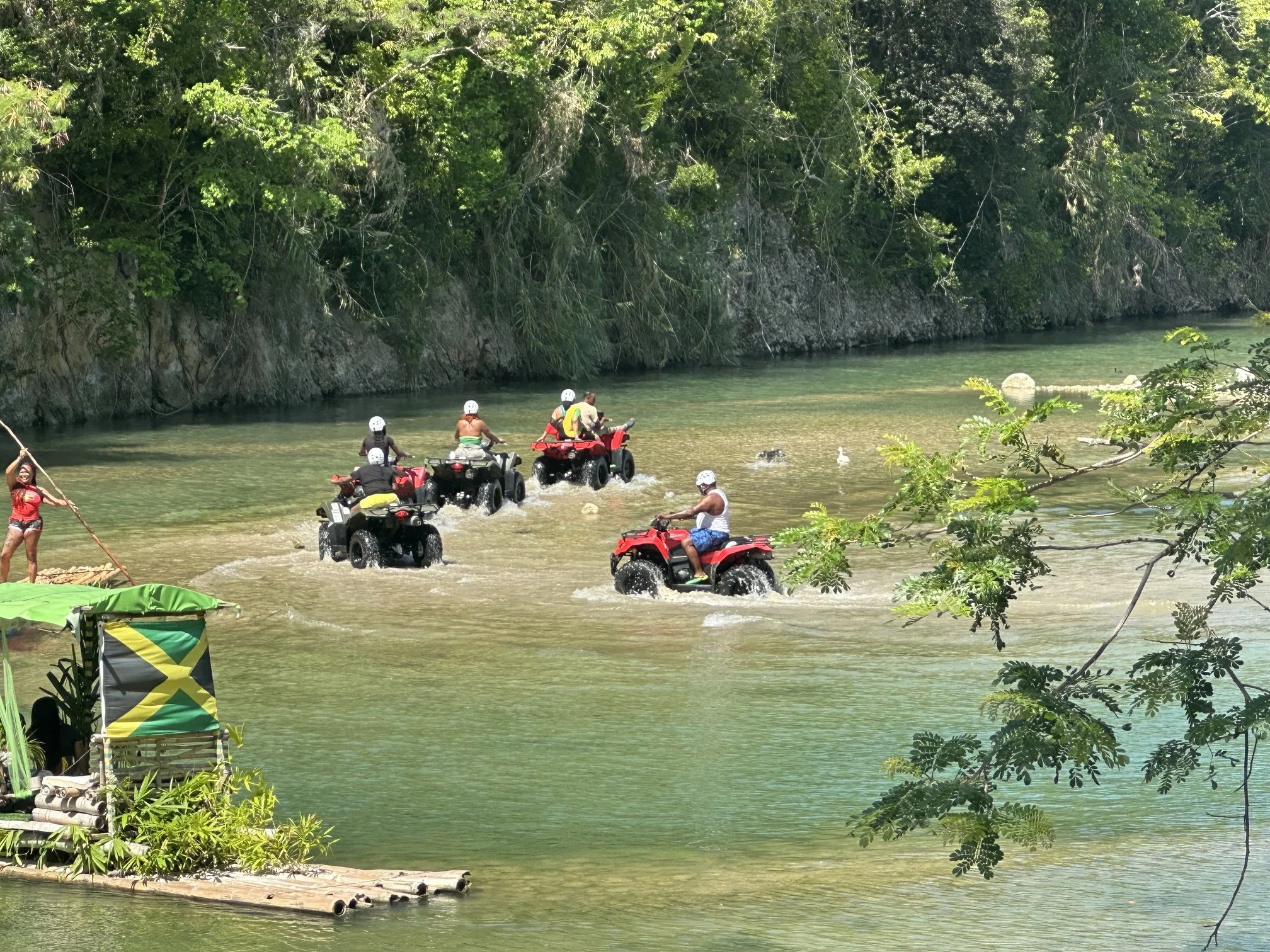 A group of people riding all-terrain vehicles (ATVs) through a shallow river surrounded by dense green forest, with some people standing on the riverbank.