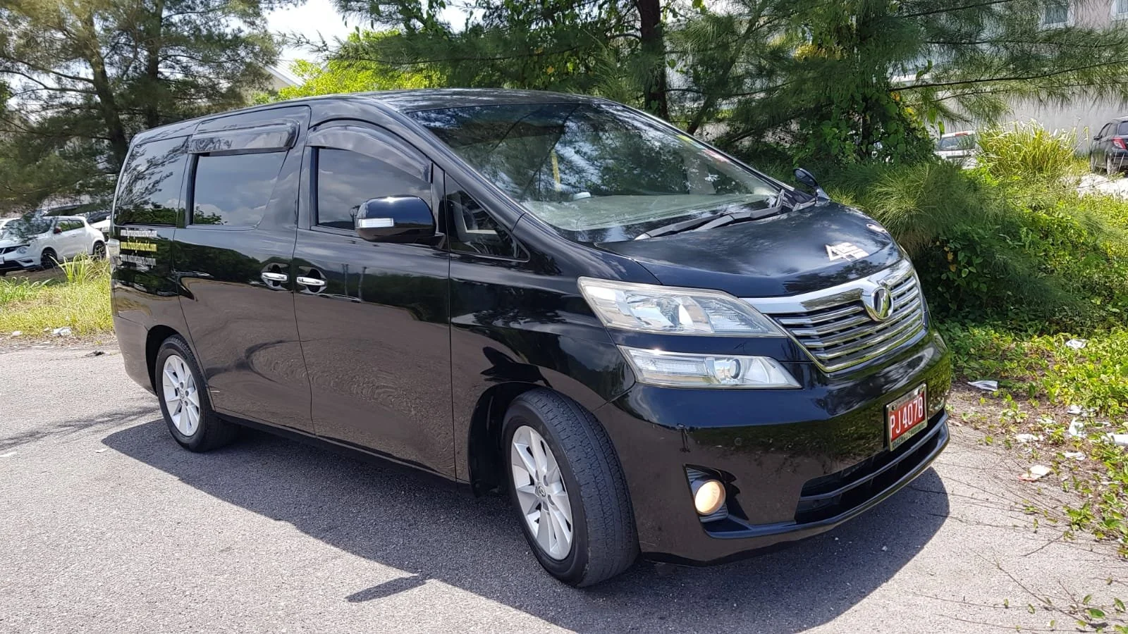 Black minivan parked on a paved area with green foliage and trees in the background.