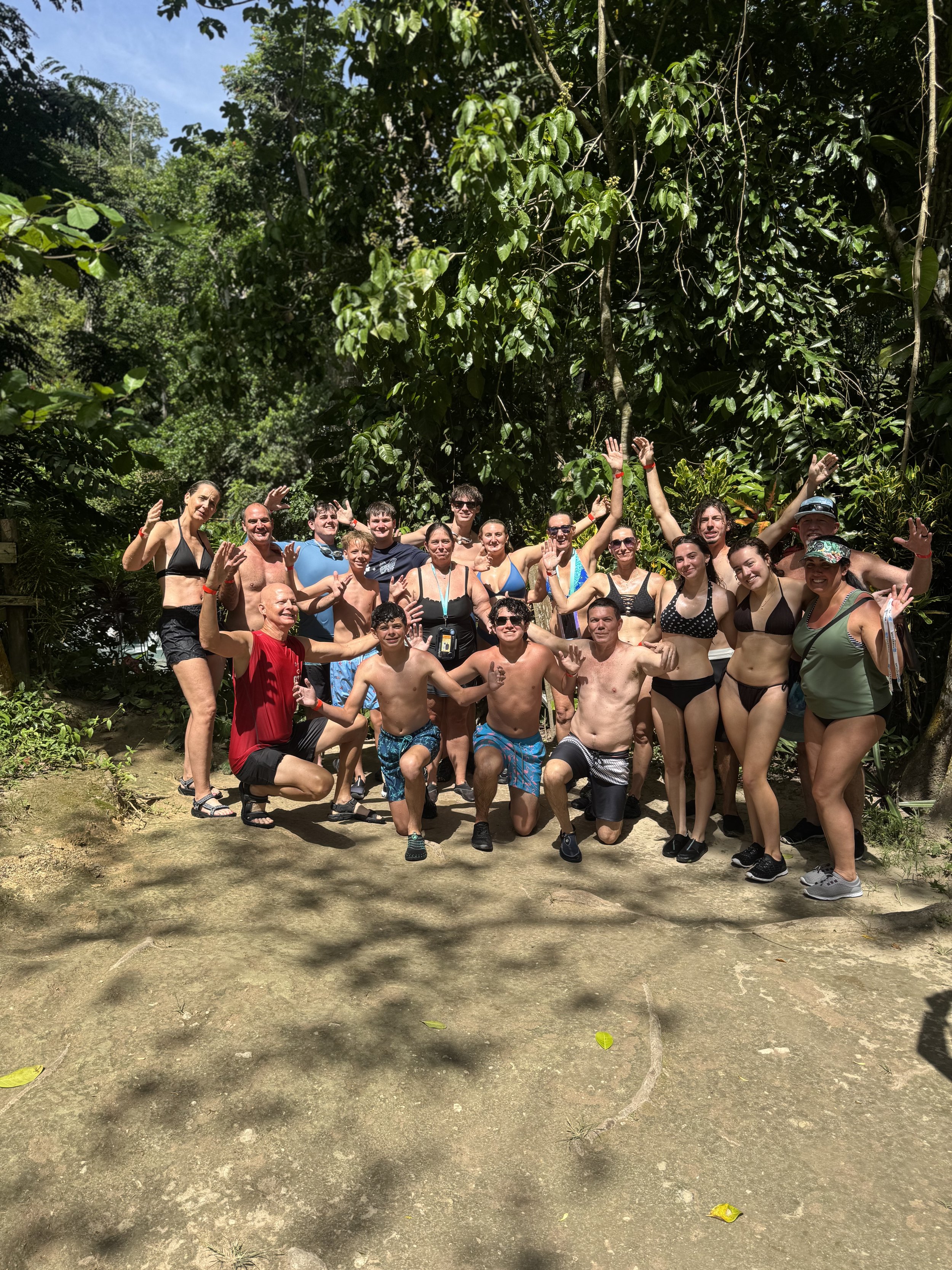 Group of around 20 people, mostly in swimwear, posing and smiling outdoors in a tropical setting with lush green trees and blue sky, some with raised hands.