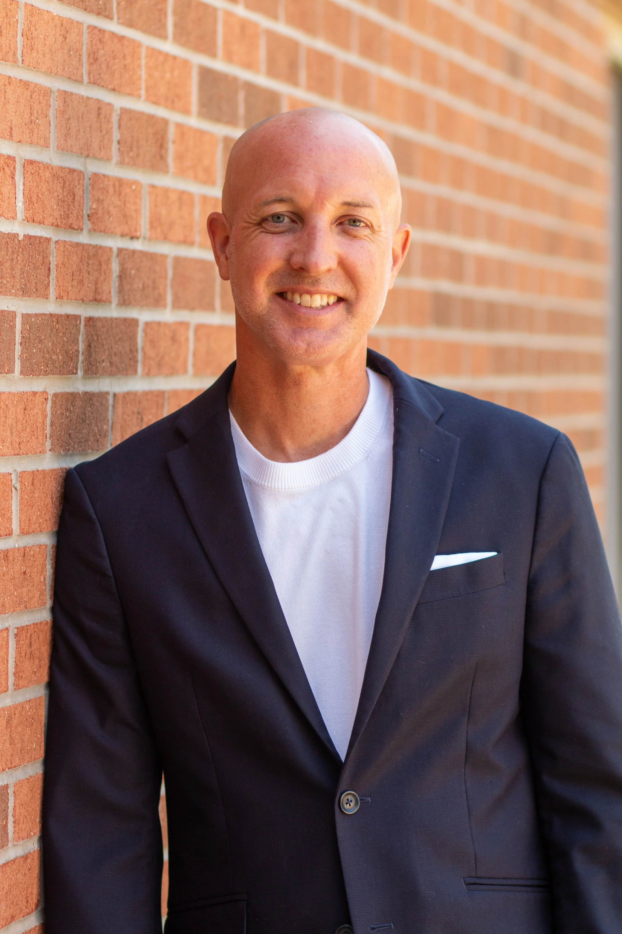 Portrait of a bald man with a friendly smile, wearing a dark blazer and white shirt, standing against a brick wall, looking at the camera.