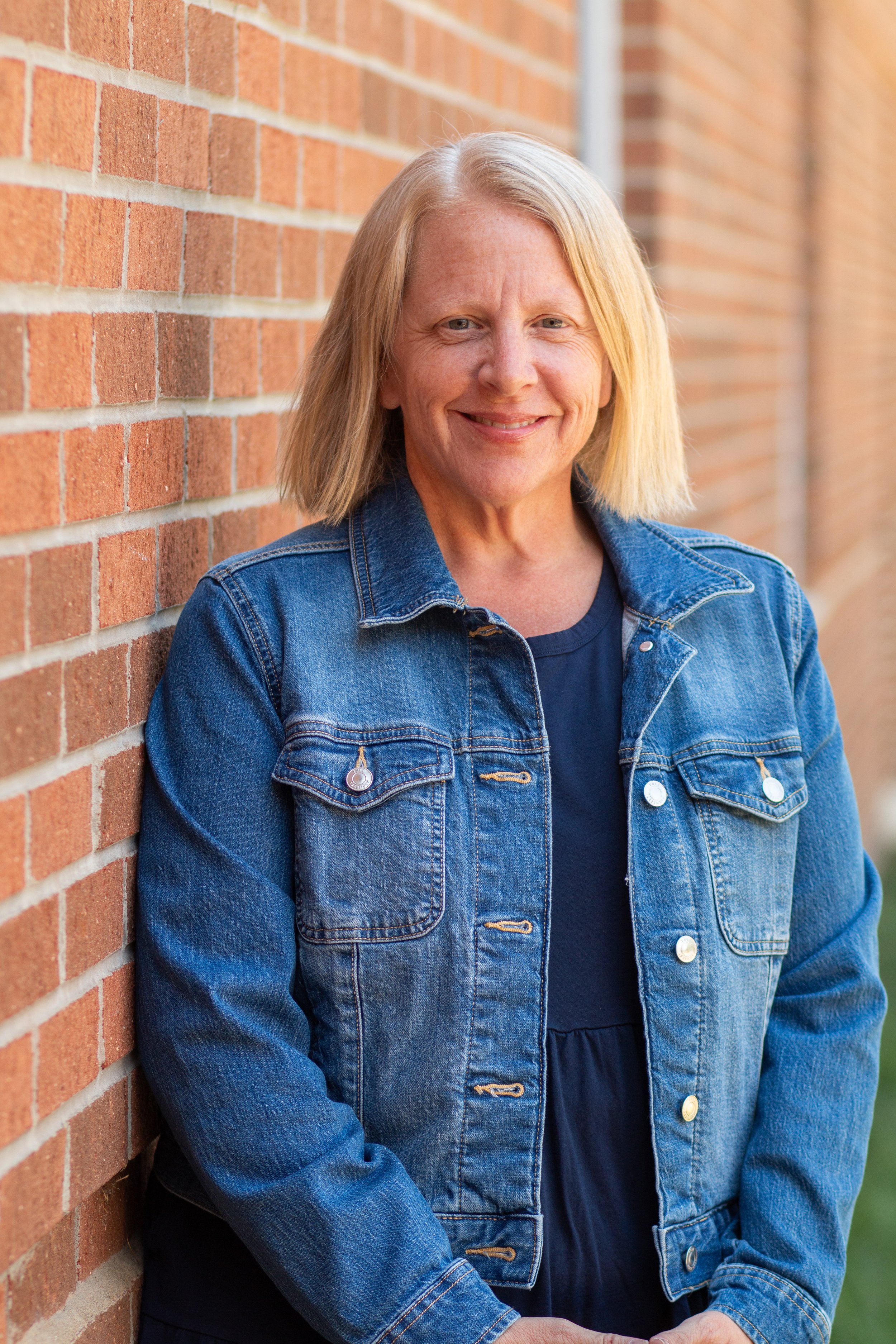 A middle-aged woman with blonde hair smiling and leaning against a brick wall outdoors during daytime.