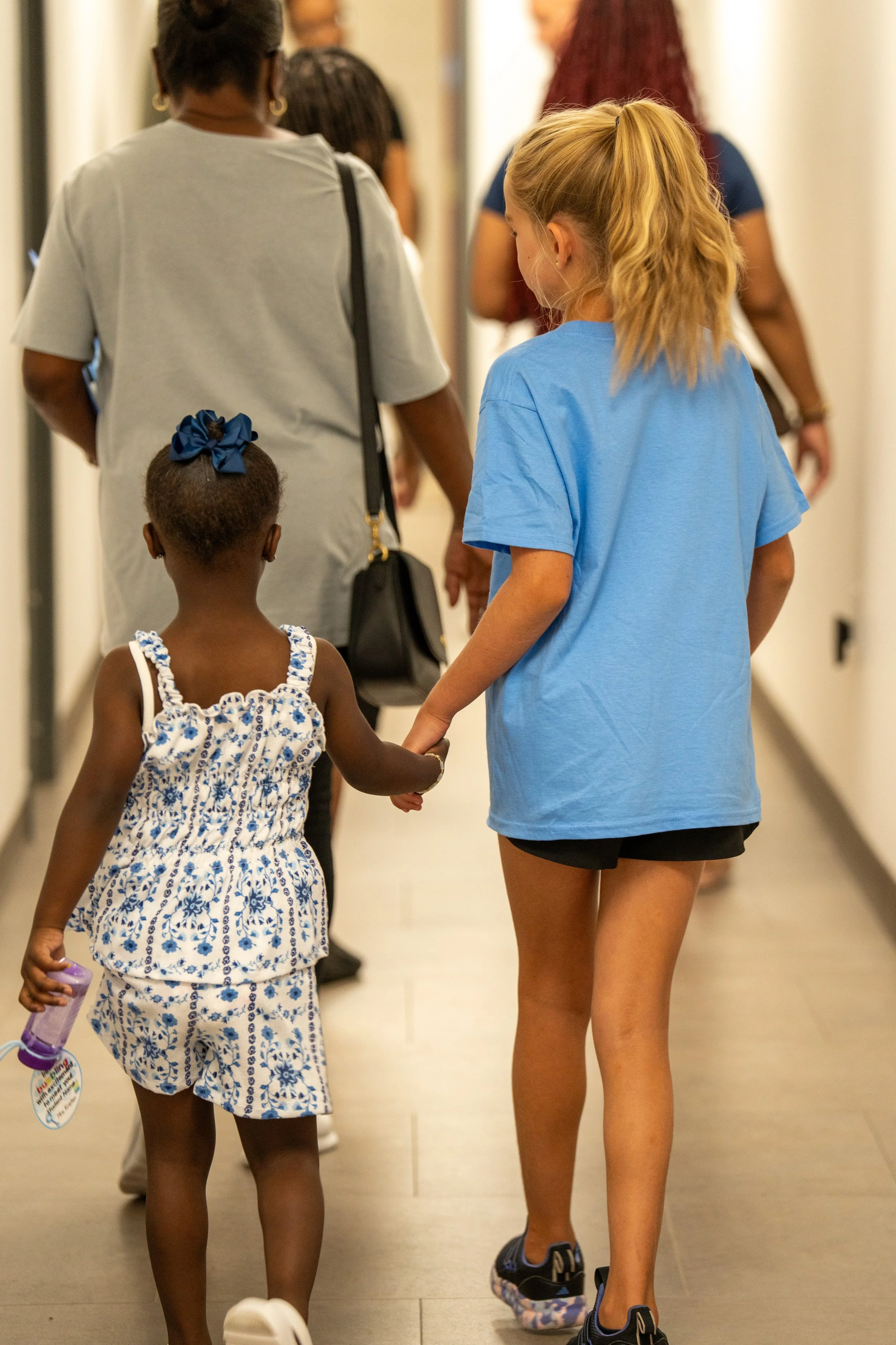 Children walking in a hallway with adults, holding hands, with one girl wearing a blue dress and a girl in a blue T-shirt and black shorts.