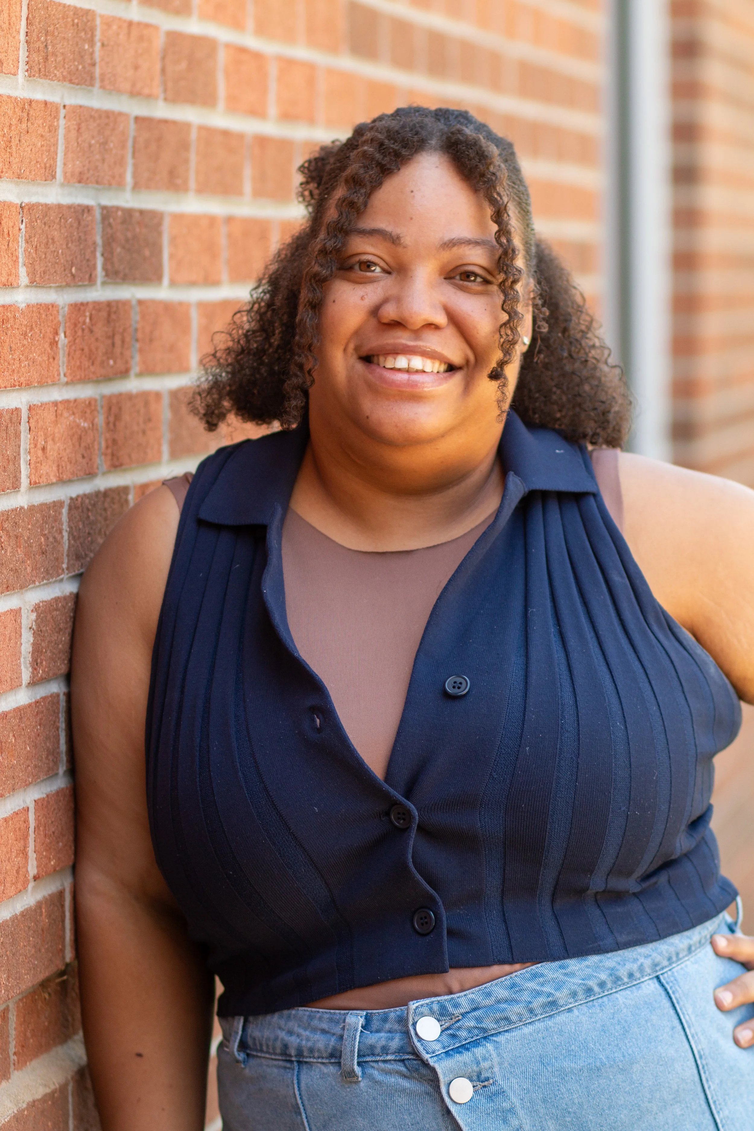 Close-up of a woman with medium skin tone, curly hair, wearing a navy sleeveless vest over a beige top, and light-wash jeans, standing against a brick wall with a smile.
