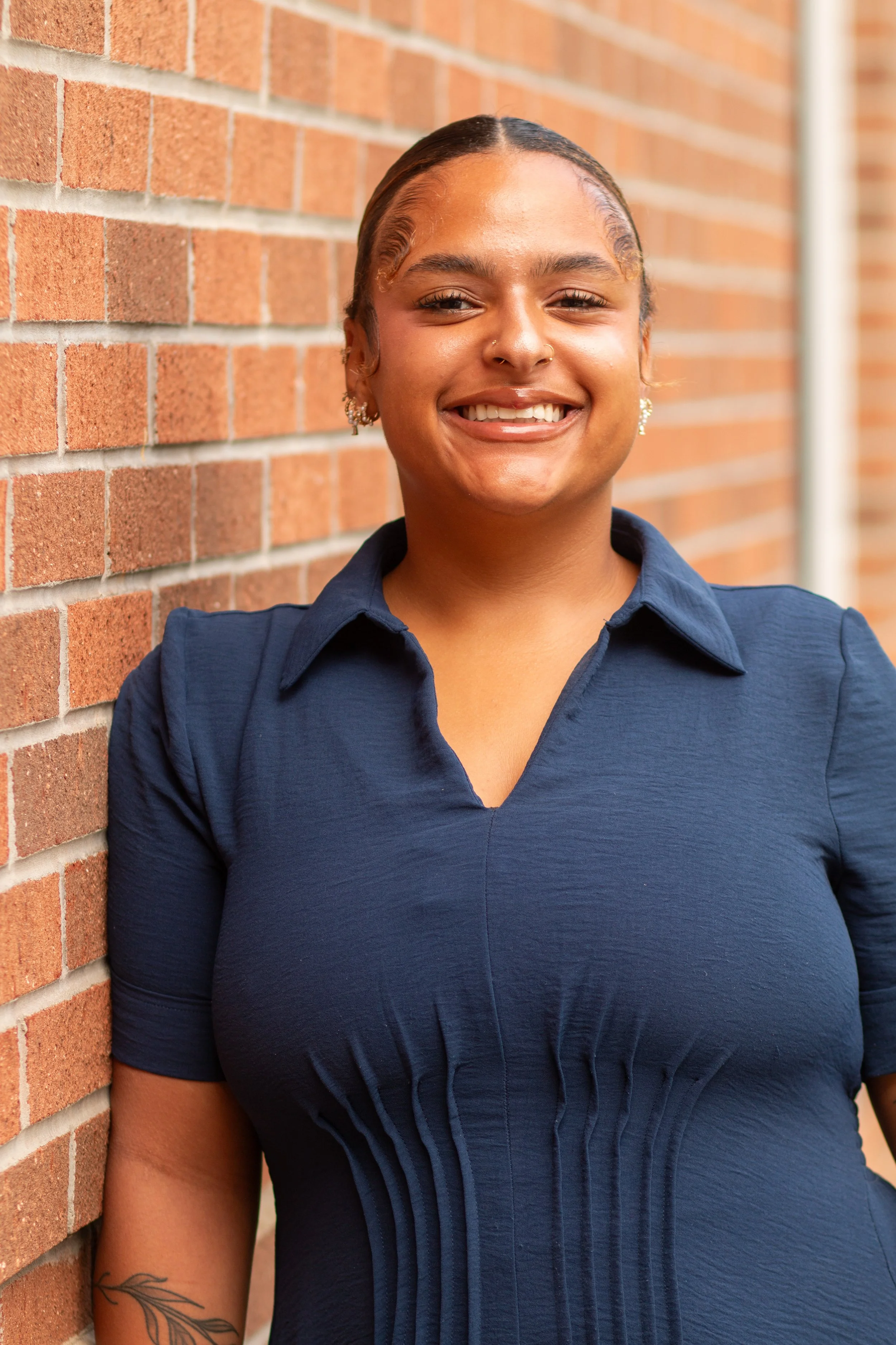 Close-up of a smiling woman leaning against a brick wall, wearing a dark blue shirt and earrings, with a turiquois nose ring and a leaf tattoo on her arm.
