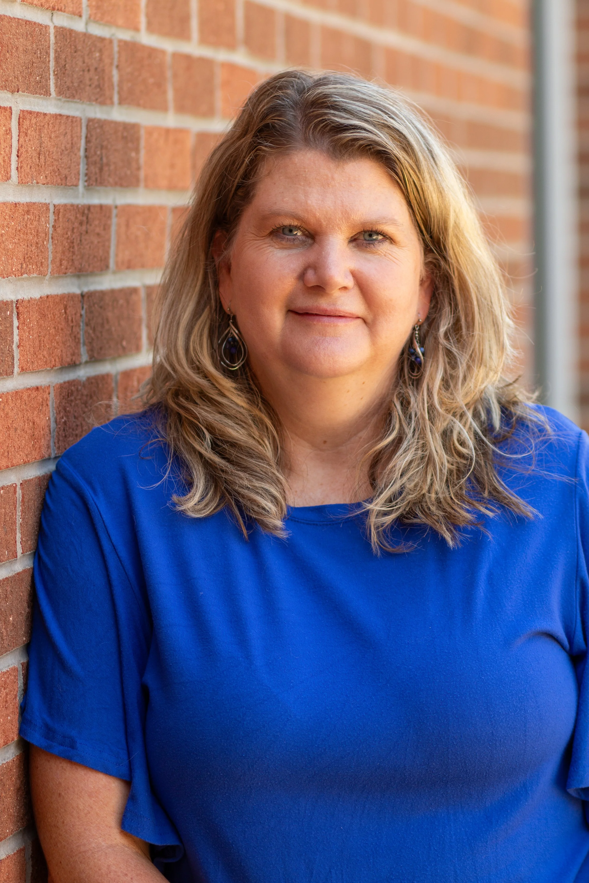 A woman with shoulder-length blonde hair and blue eyes, wearing a royal blue top, stands in front of a brick wall. She is looking at the camera with a slight smile.