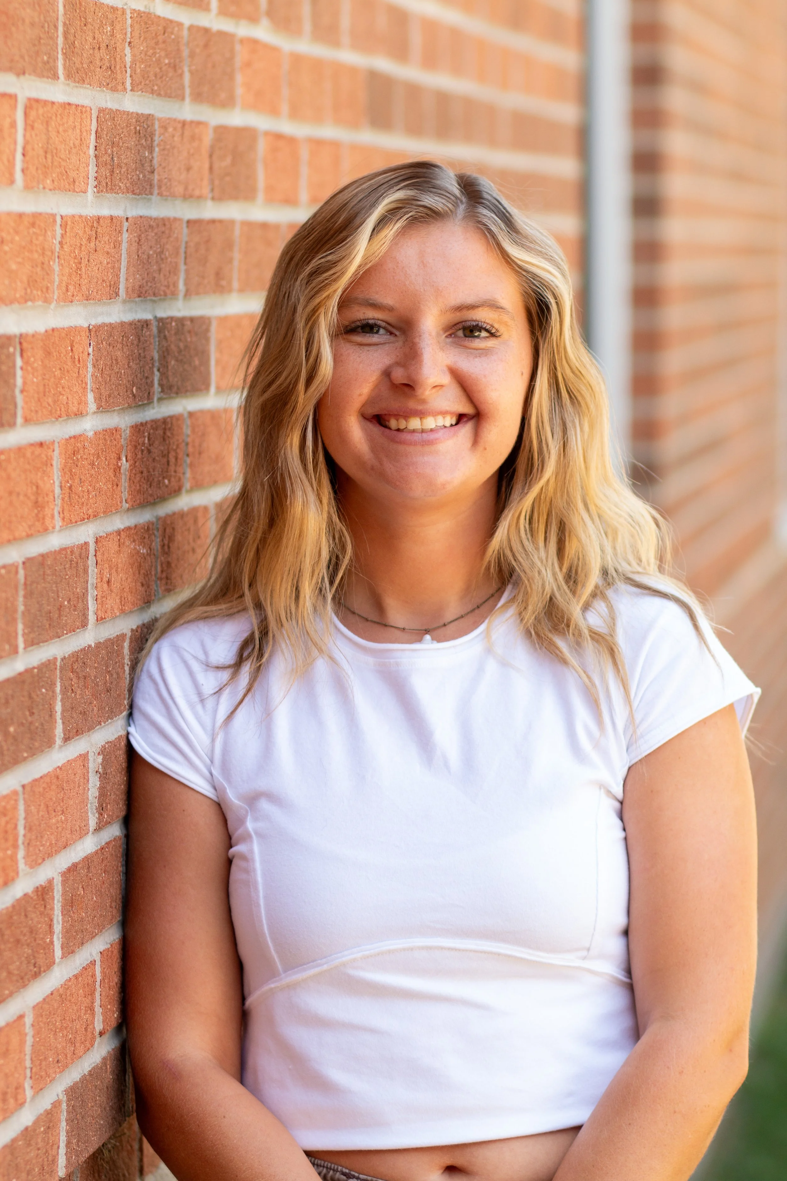 A young woman with wavy blonde hair and a white T-shirt, smiling while leaning against a brick wall outdoors.