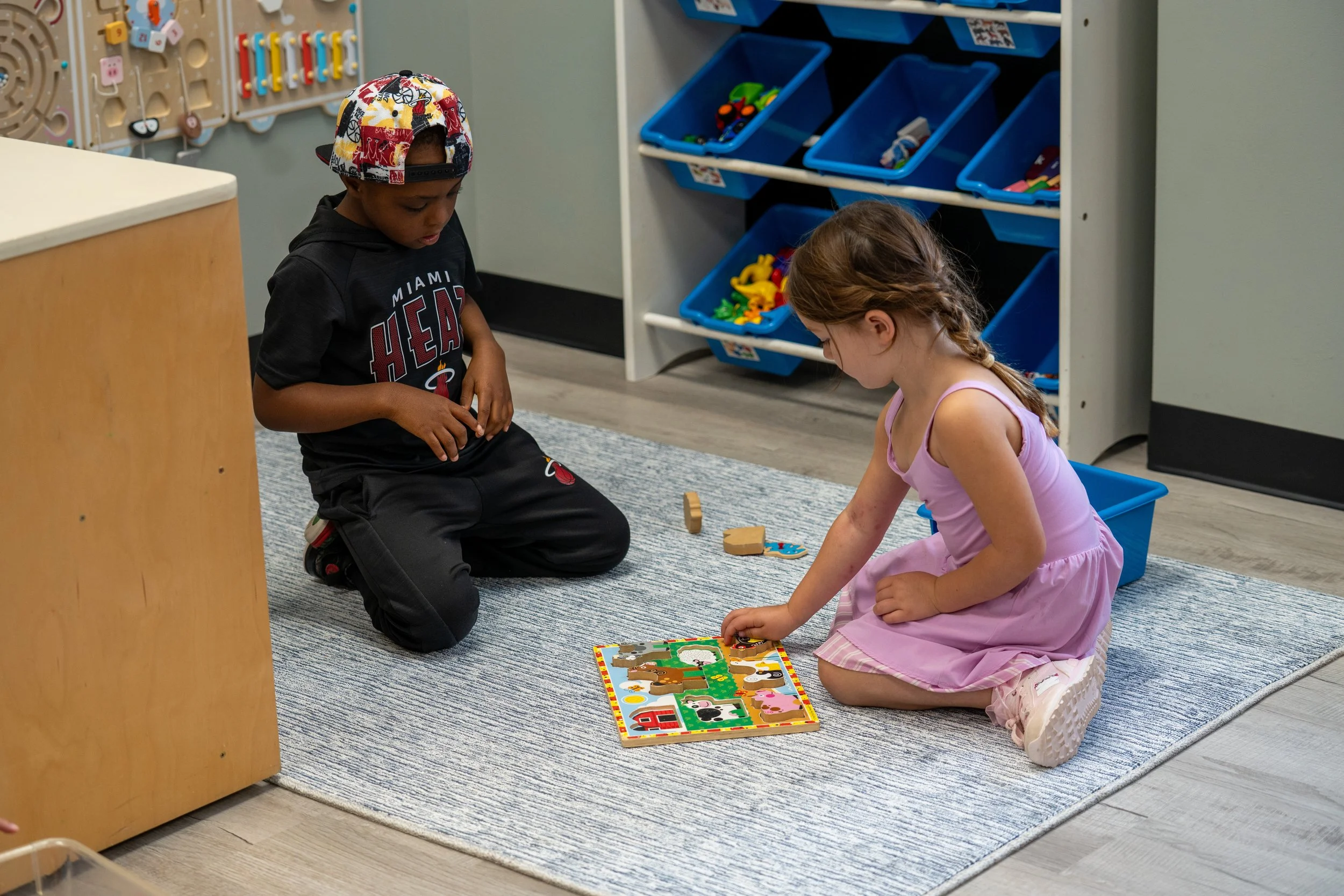 Two children, a boy and a girl, sitting on the floor playing with a colorful puzzle in a classroom with shelves of toys around them.