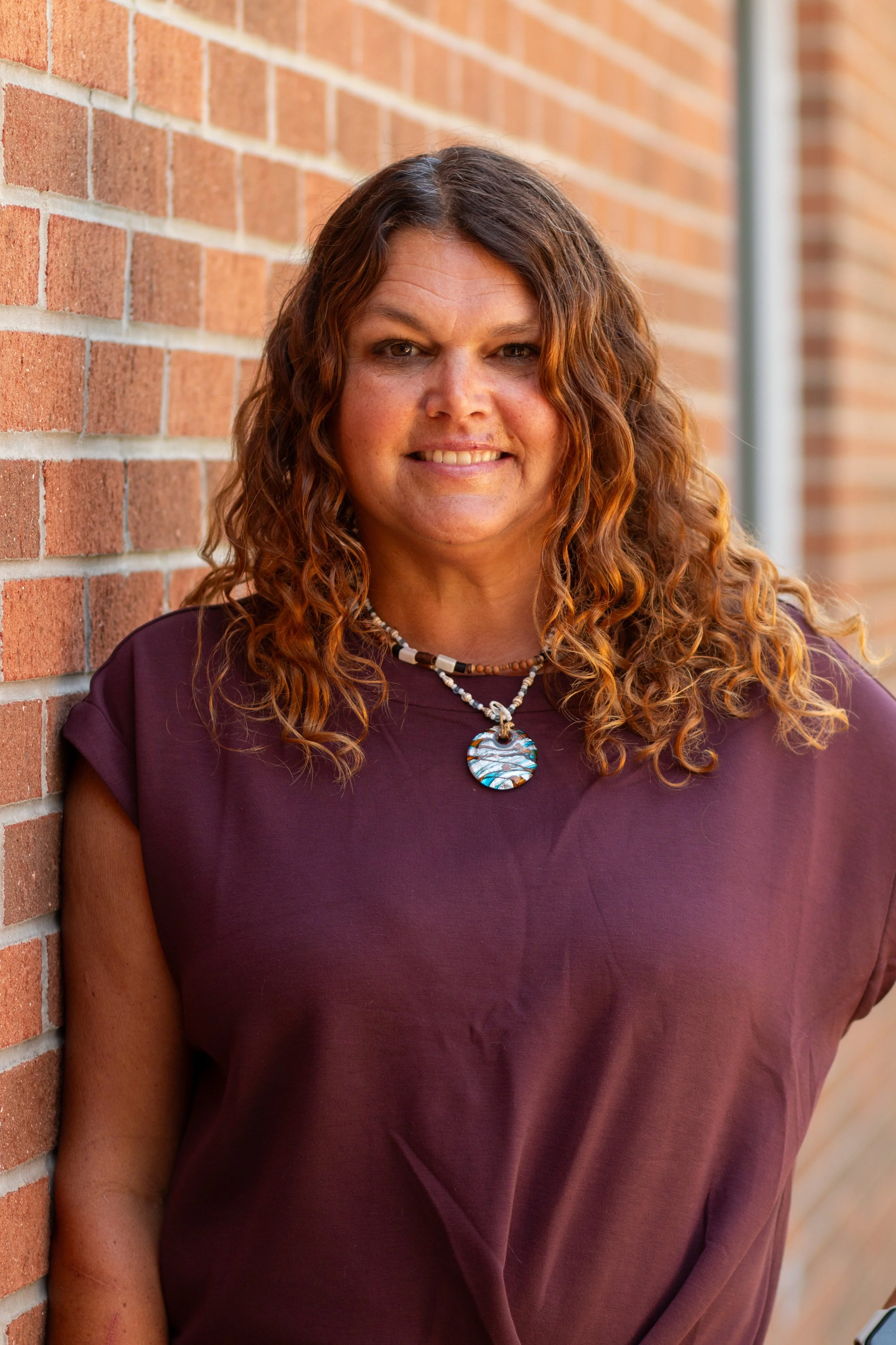 A woman with curly reddish-brown hair smiling, standing against a brick wall, wearing a maroon top and beaded necklaces.